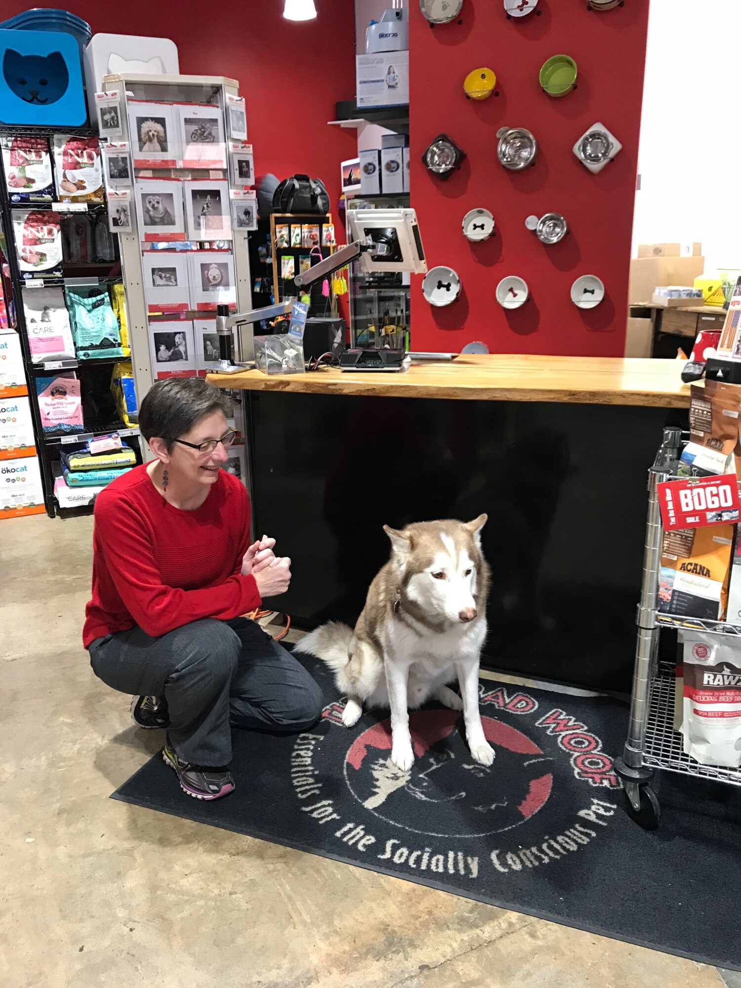 Person crouching next to a dog inside a store with shelves and a red wall display behind them.
