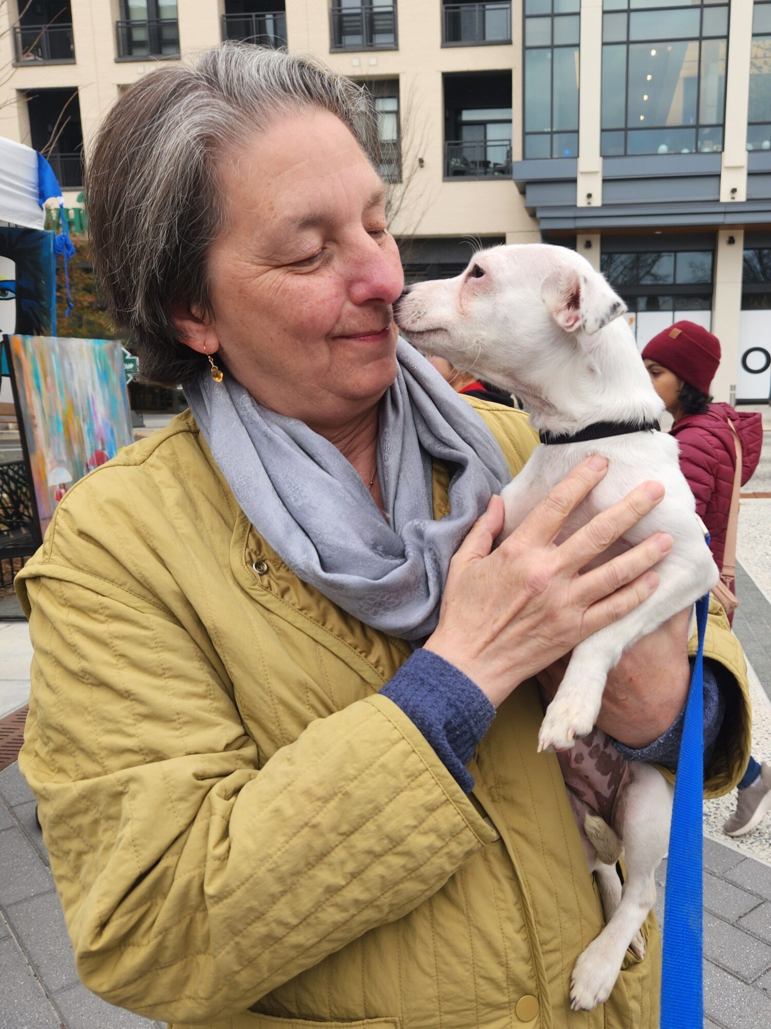 Woman holding a small white dog close, outdoors in an urban setting, smiling with eyes closed.