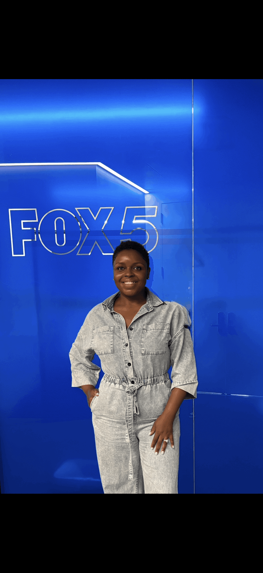 Woman smiling in front of a blue FOX 5 backdrop, wearing a light-colored jumpsuit.