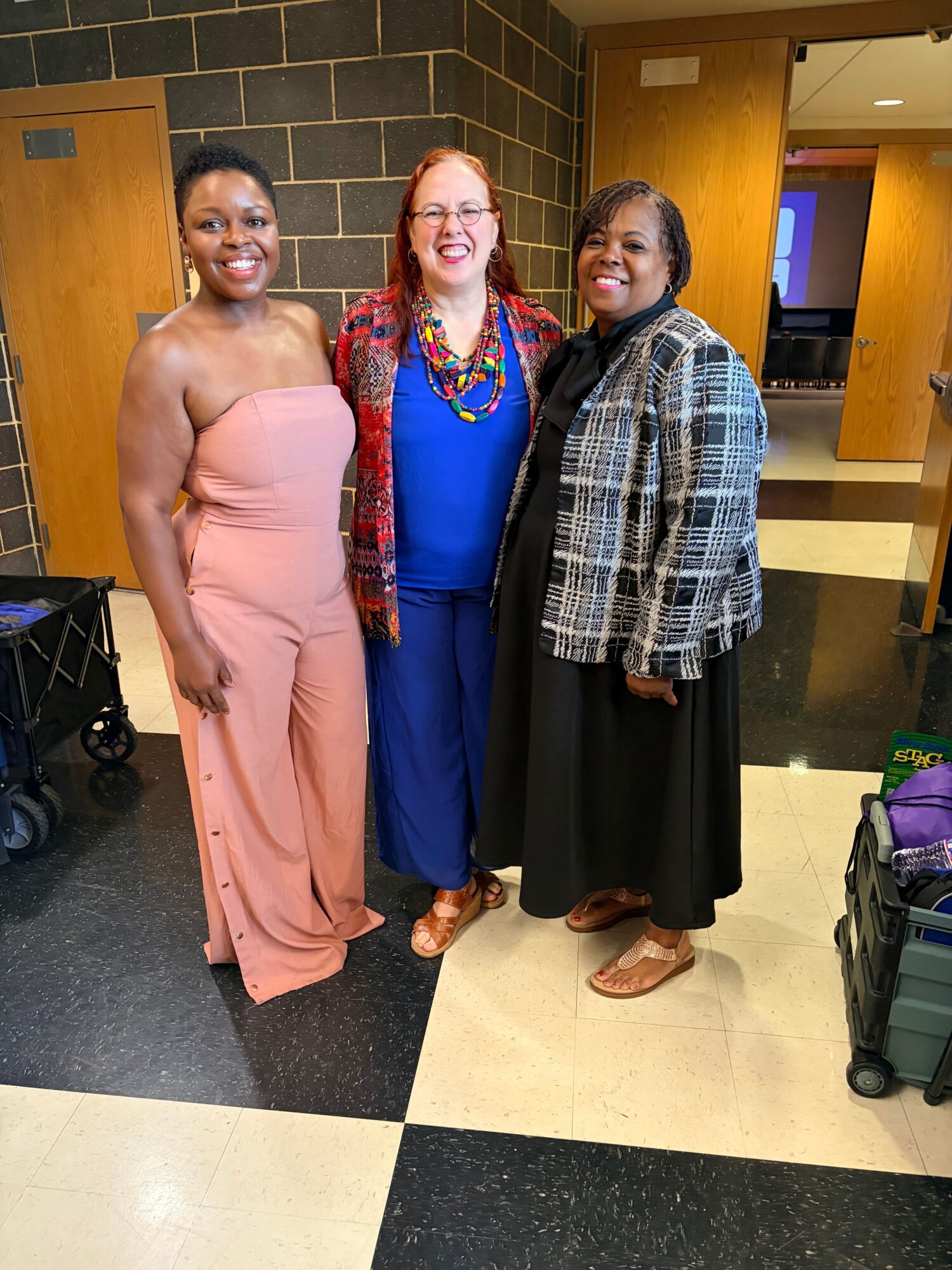 Three women standing indoors, one in a plaid jacket, one in a colorful dress, and one in a pink dress, smiling.