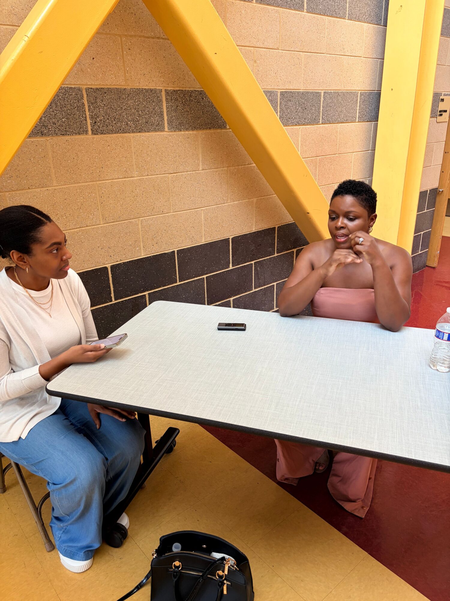 Two women sitting at a table, one with a phone, in a room with yellow and brown walls.