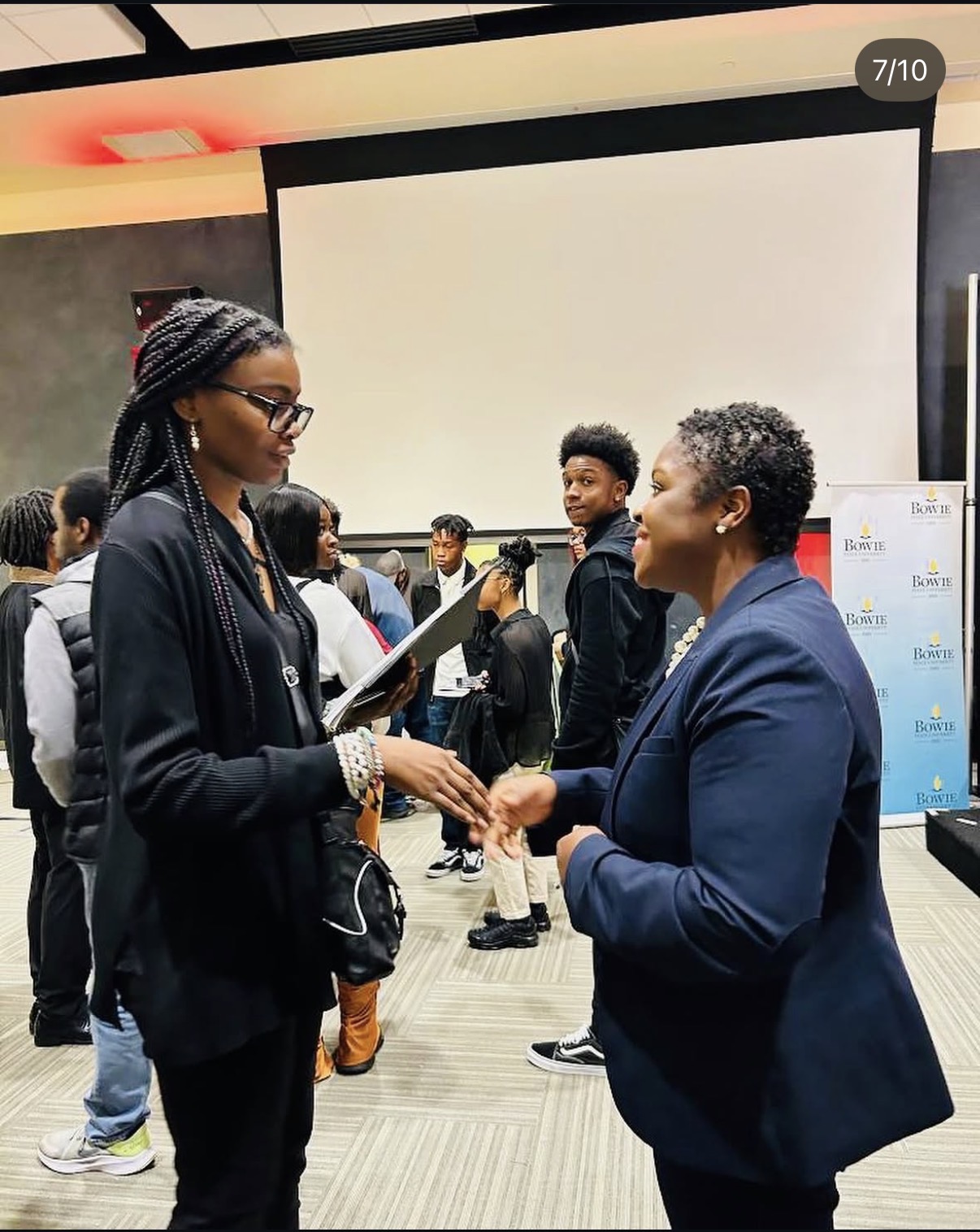 Two women shaking hands in a conference room with a large screen behind them, surrounded by people. One woman wears glasses and a black jacket, the other wears a blue blazer.