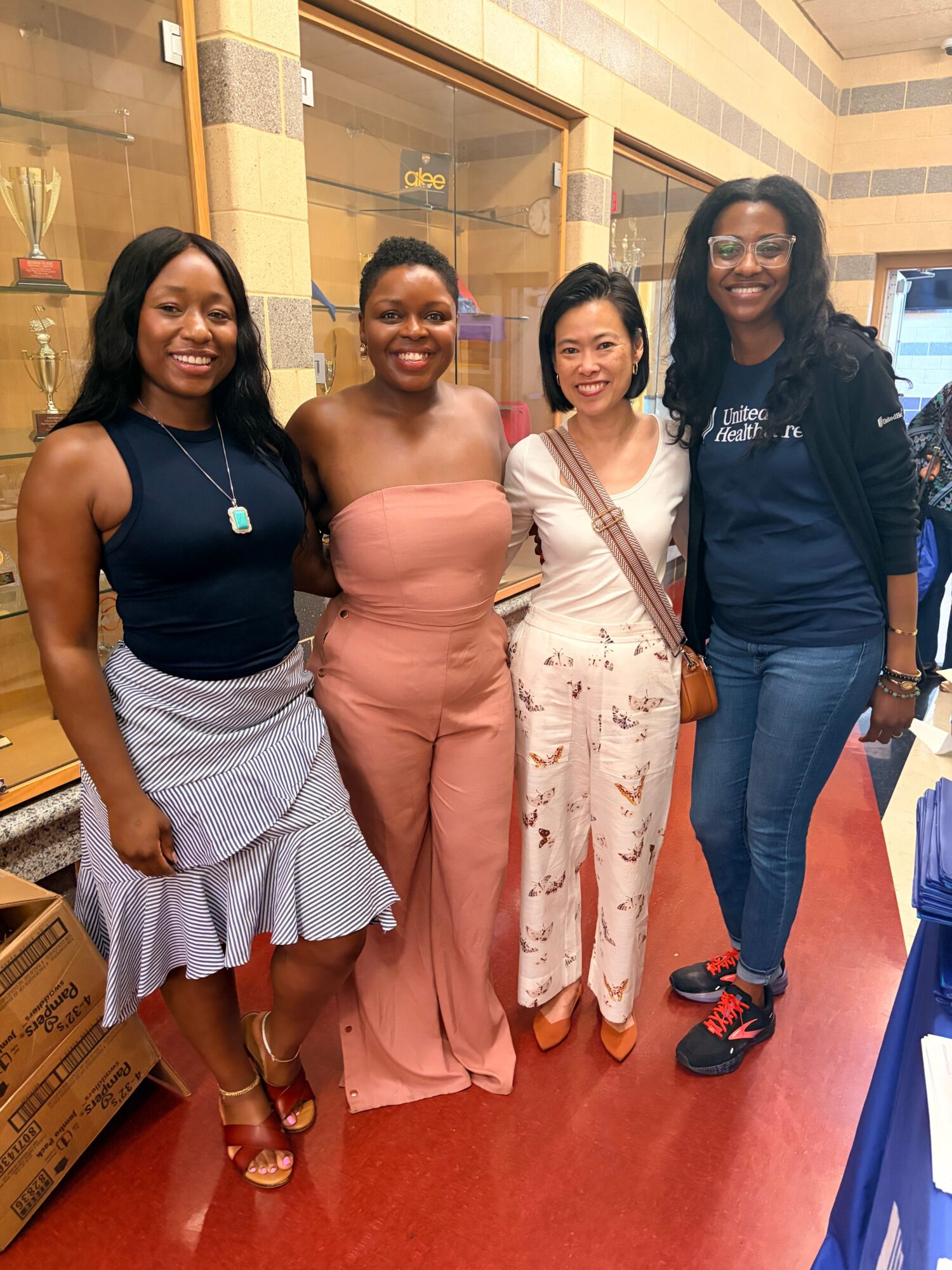 Four women standing indoors on a red carpet, smiling, with a glass door and yellow walls in the background.