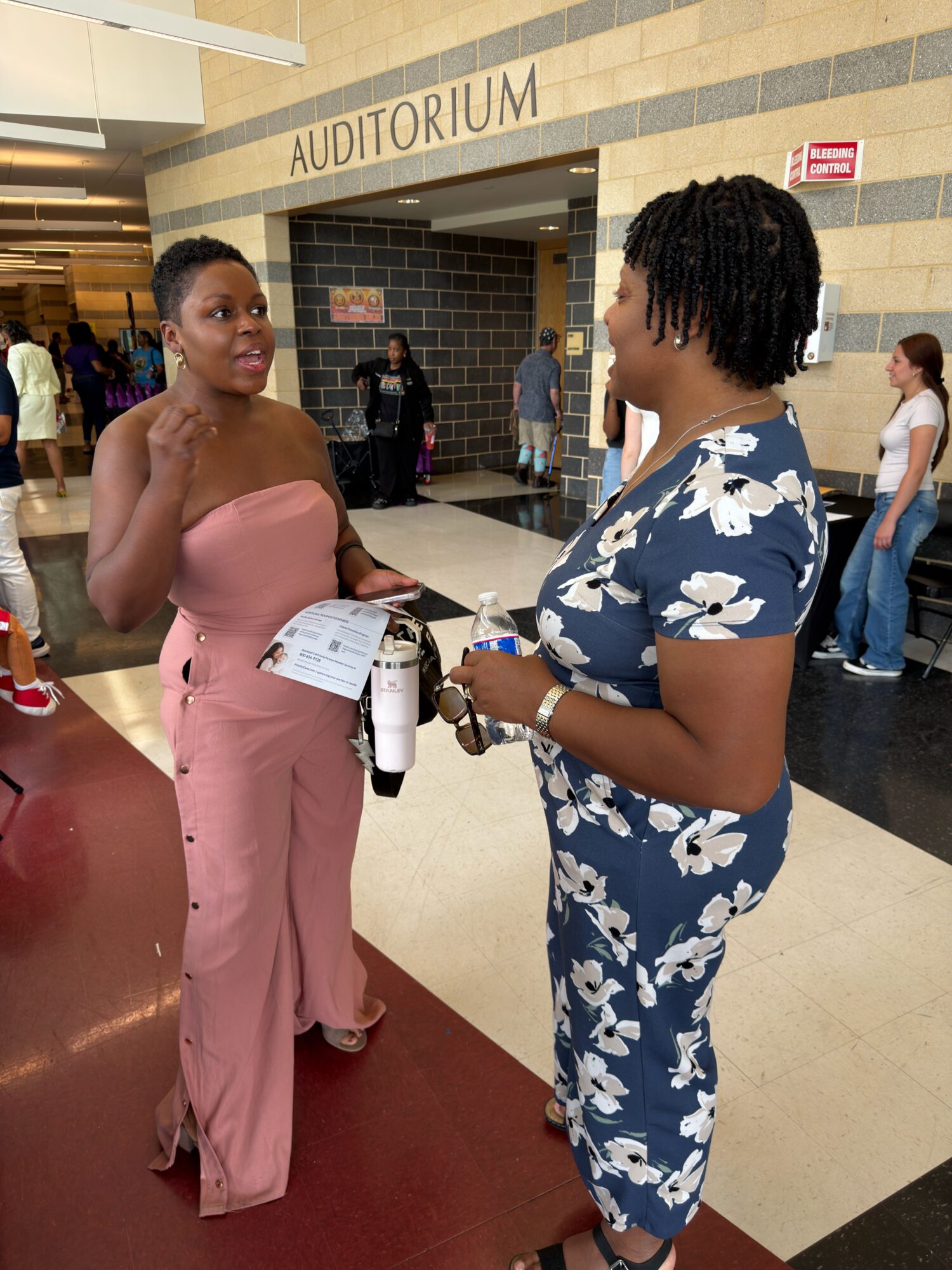 Two women in a hallway, one in a floral jumpsuit and the other in pink pants, holding a bottle and a paper, smiling.