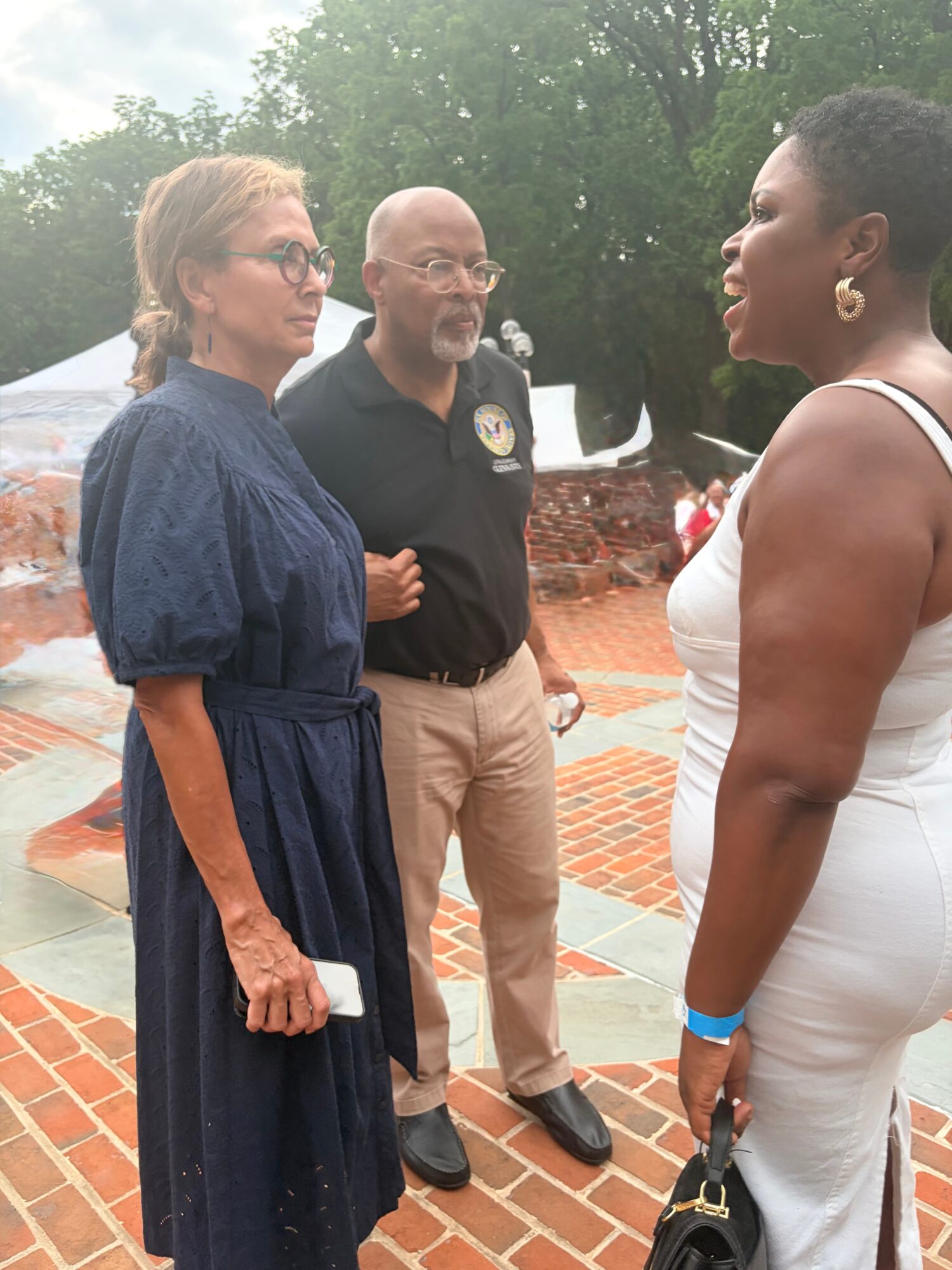 Three people engaged in conversation outdoors, with trees and a brick structure in the background.