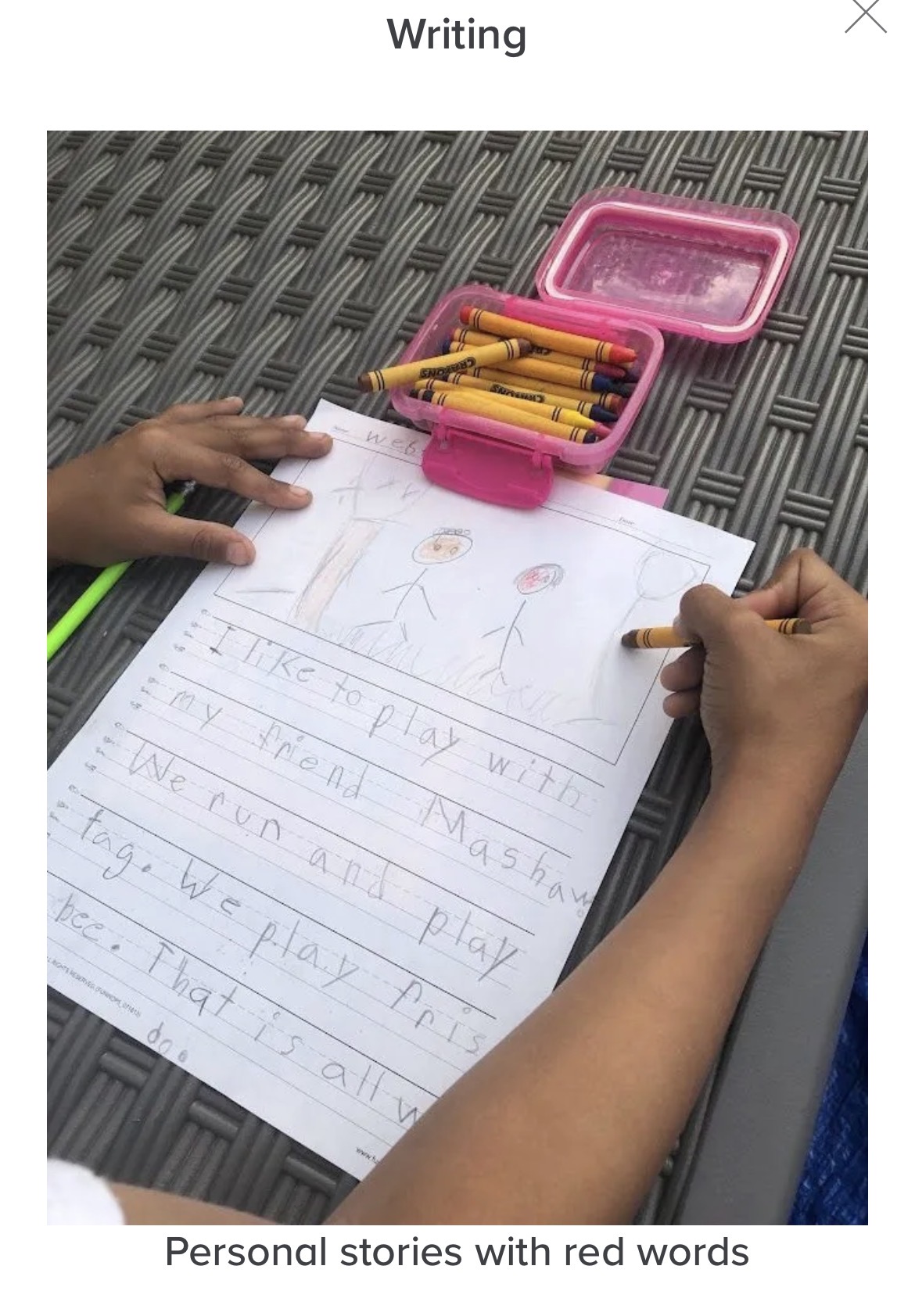 Child's hand writing on paper with a pink pencil case and colored pencils on a textured surface.