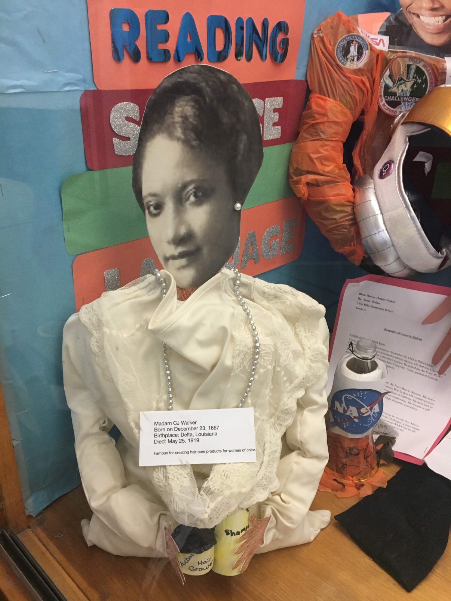 A paper doll with a black-and-white photo of a person's face, dressed in a white outfit, lying on a table with various objects around.