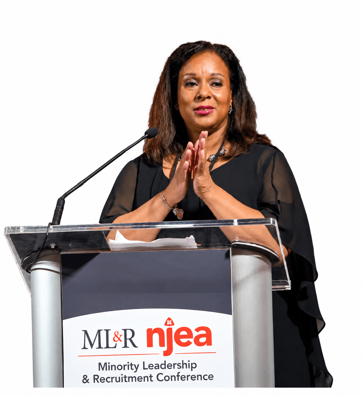 Woman standing at a podium with hands clasped, speaking at a conference, with a logo and text on the podium.