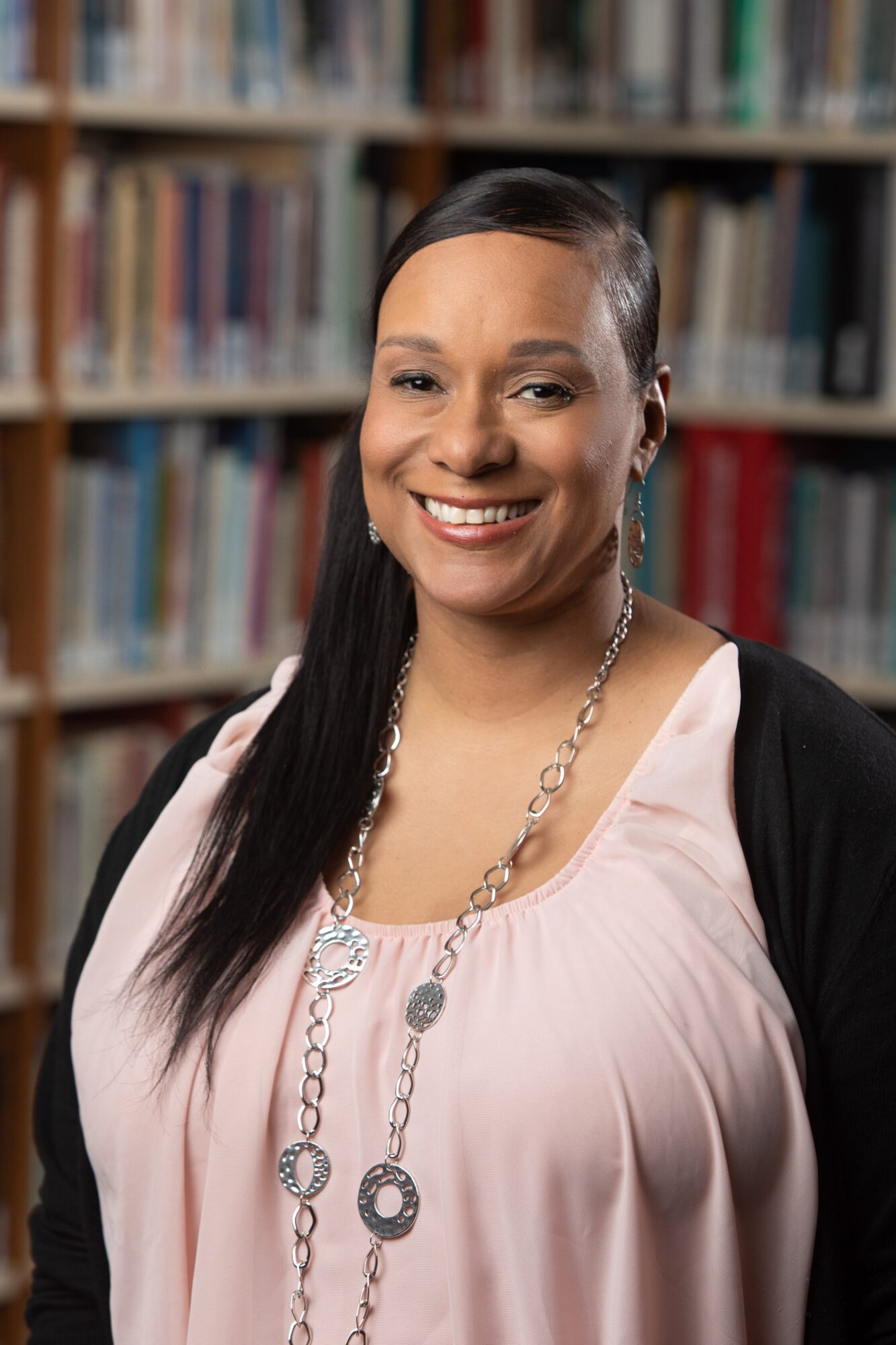 Smiling woman with long dark hair and earrings in a library, wearing a light pink top and necklace.