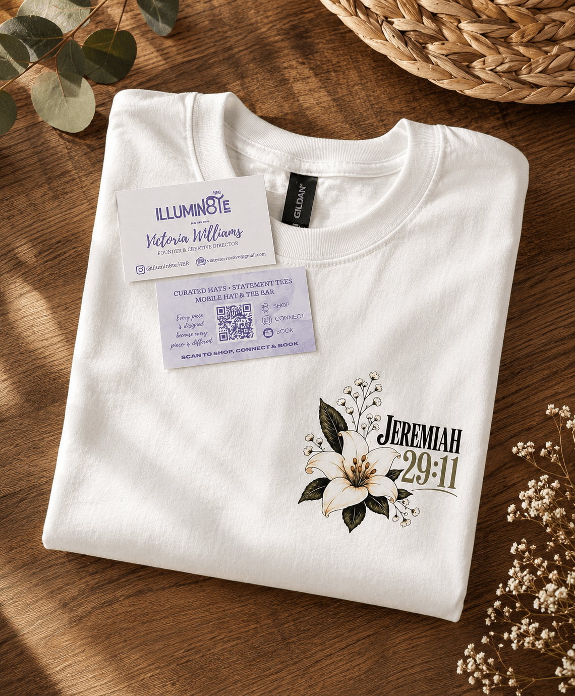 White T-shirt with floral design and text, placed on a wooden surface with a woven basket and flowers nearby.