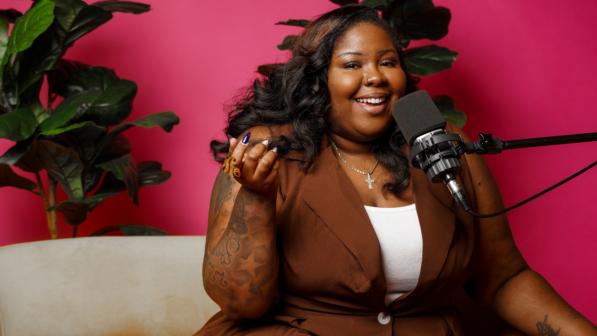 Smiling woman with long dark hair, wearing a brown top, speaking into a microphone against a pink background with green plants.