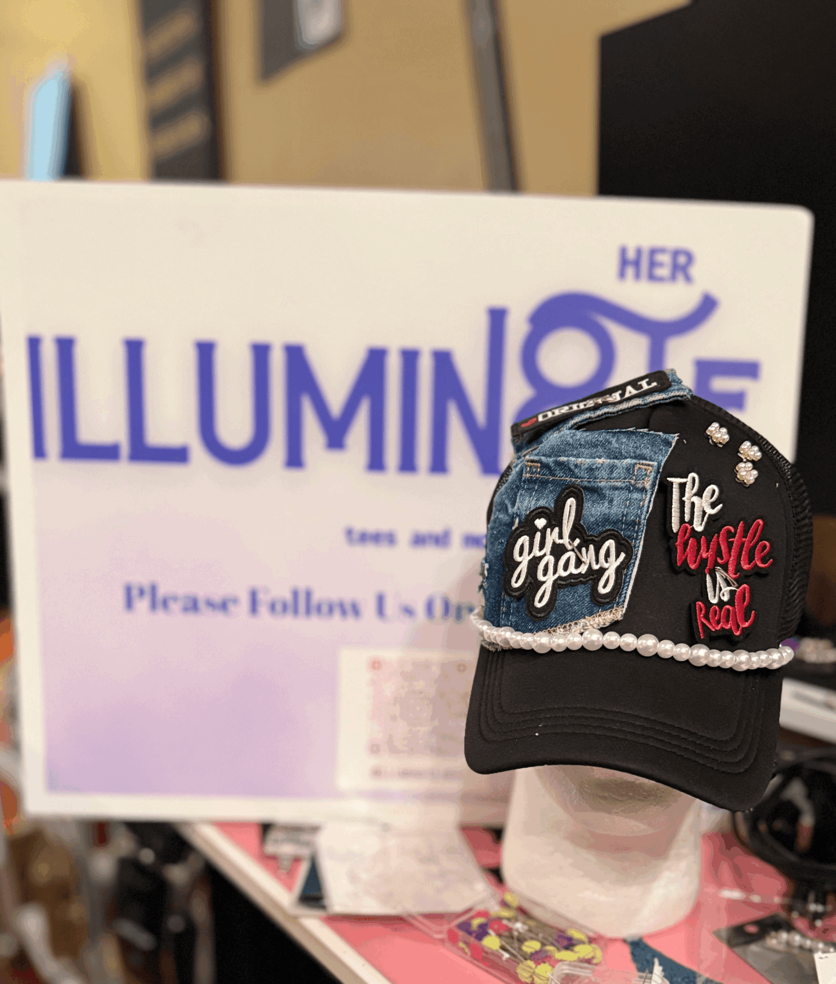 Cap with girl gang and hustle love patches displayed on a mannequin head in front of a sign. Background includes a desk and office items.