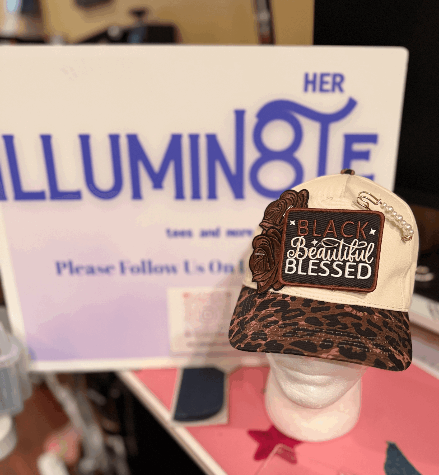 Cap with a patch reading 'Black, Beautiful, Blessed' on a mannequin head, in front of a sign with large purple text.