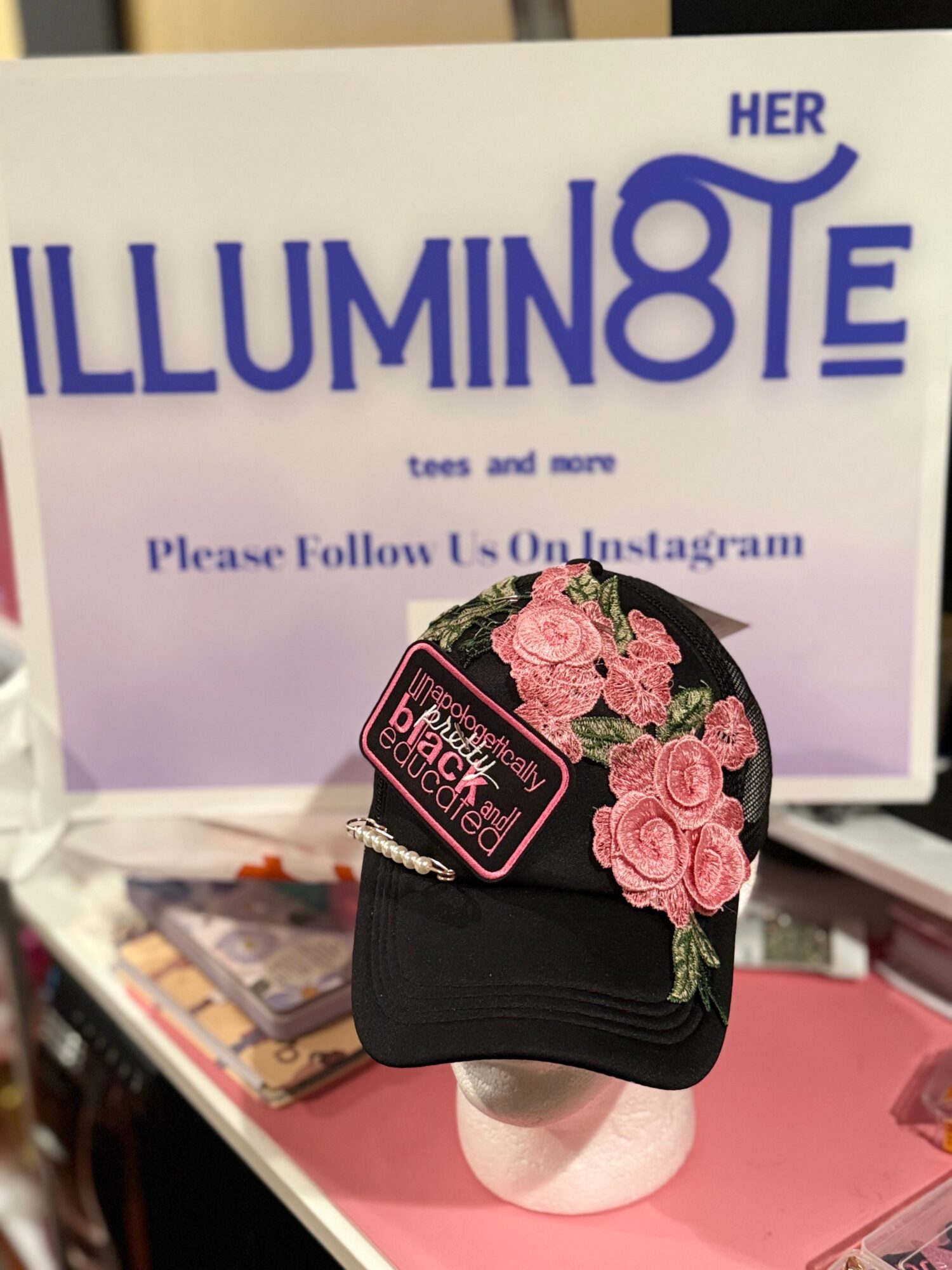 Black cap with pink roses and a pink tag, displayed on a white stand in front of a sign. The sign has blue text.