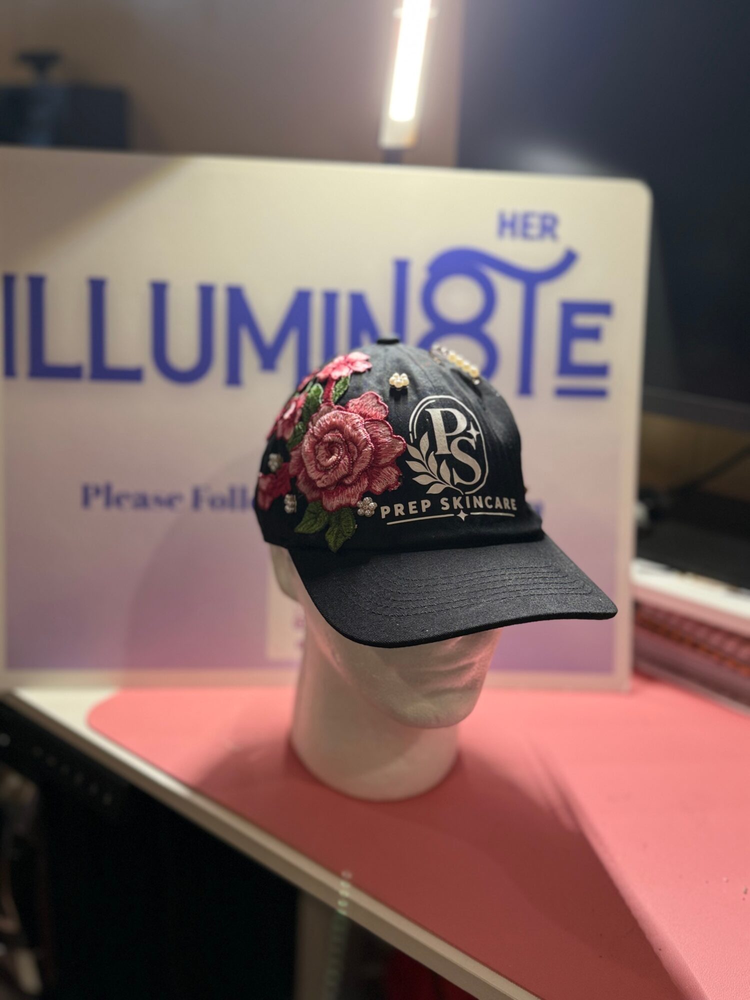 Black cap with floral embroidery on a mannequin head, displayed in front of a sign with partially visible text, on a pink surface.