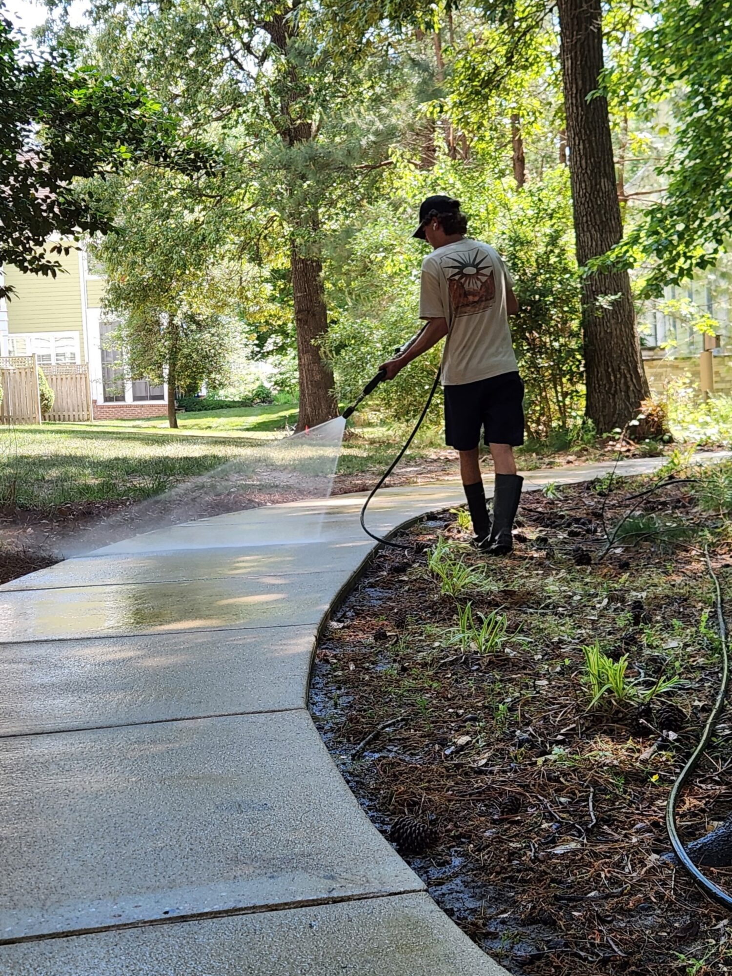 Person watering a sidewalk with a garden hose in a park with trees and grass, sunlight filtering through leaves.