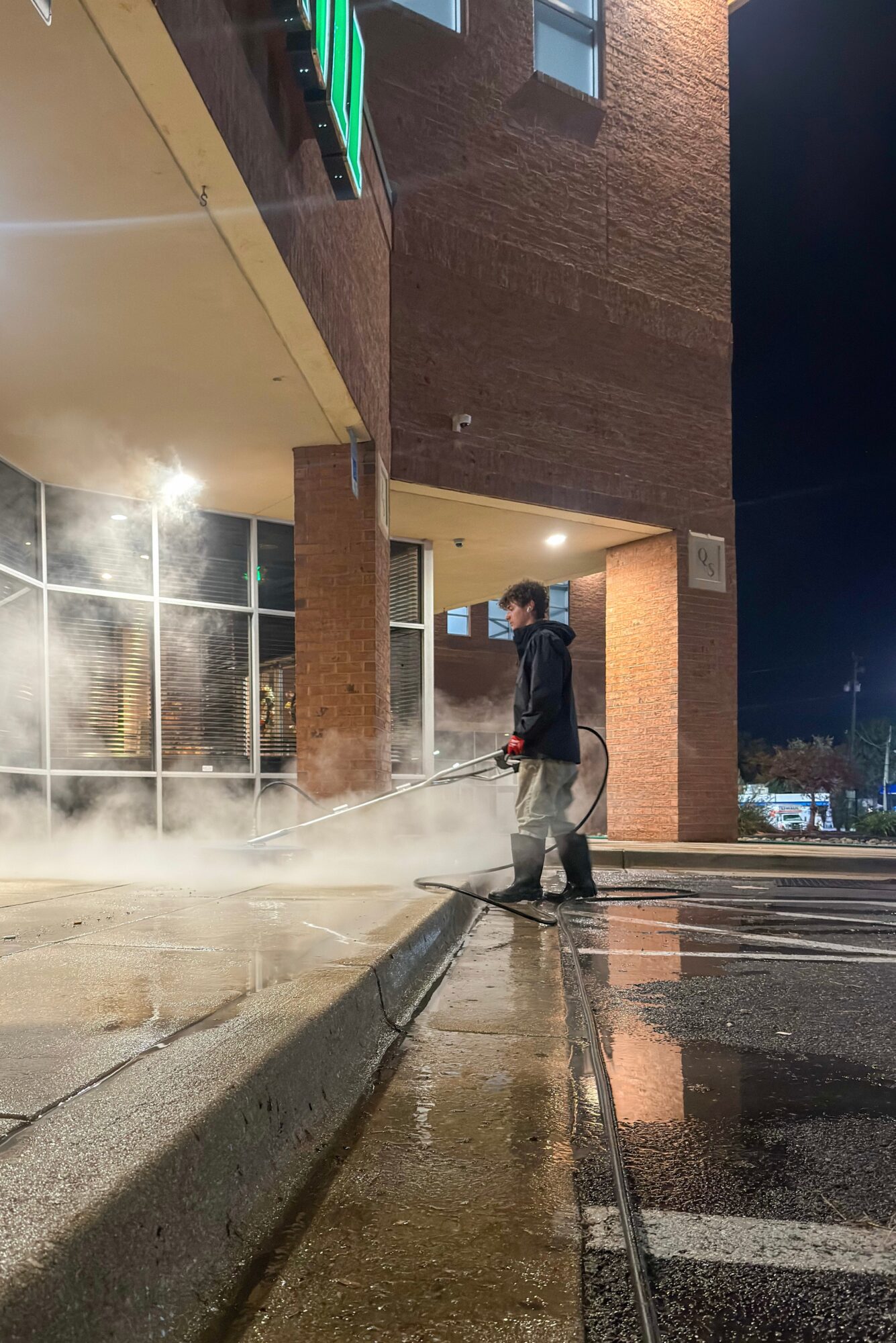 Person cleaning the sidewalk outside a building at night, steam rising from the ground, wet surface reflecting lights.