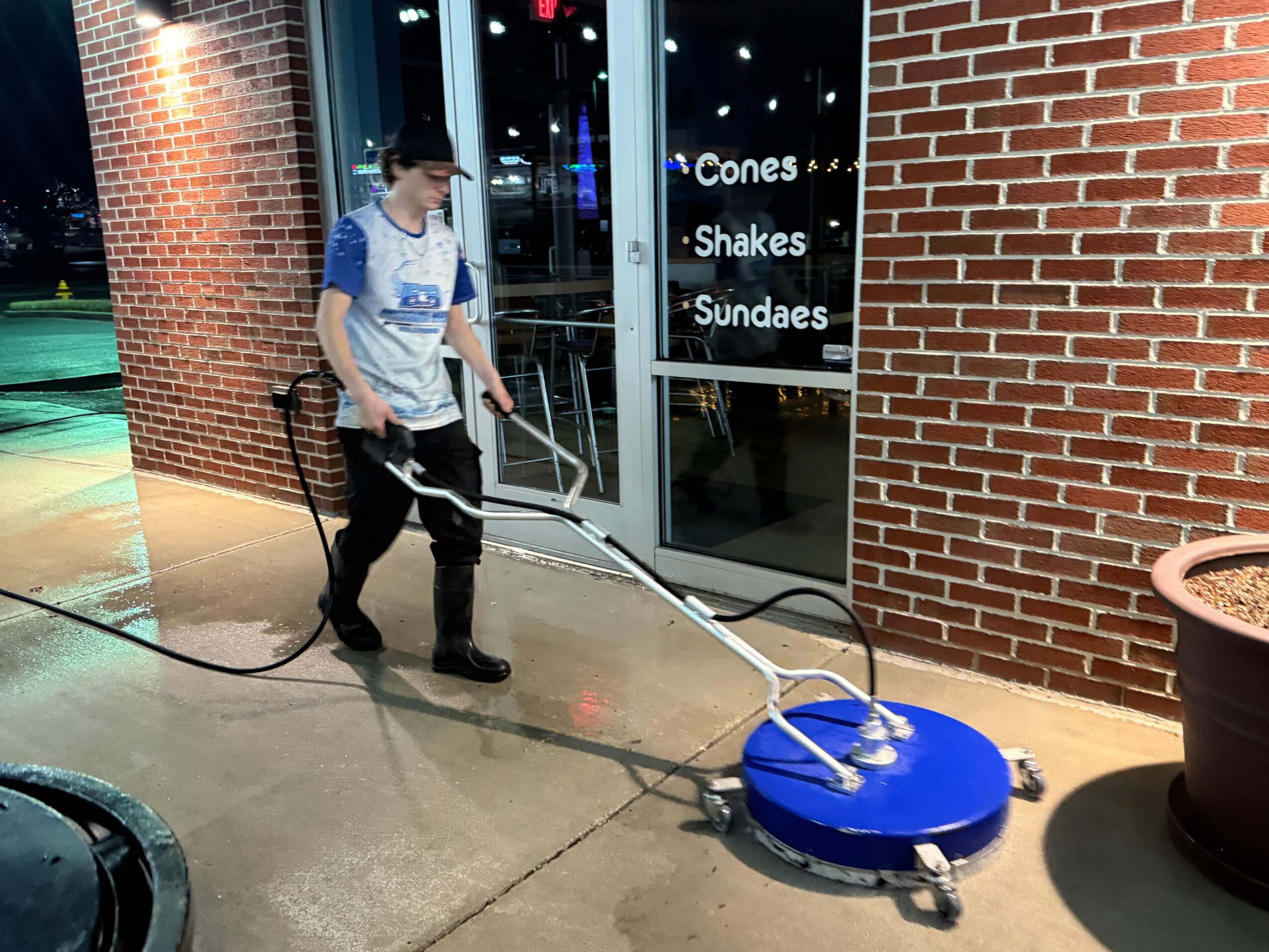 Person cleaning a window with a blue cleaning tool, brick wall, and illuminated signs in the background.