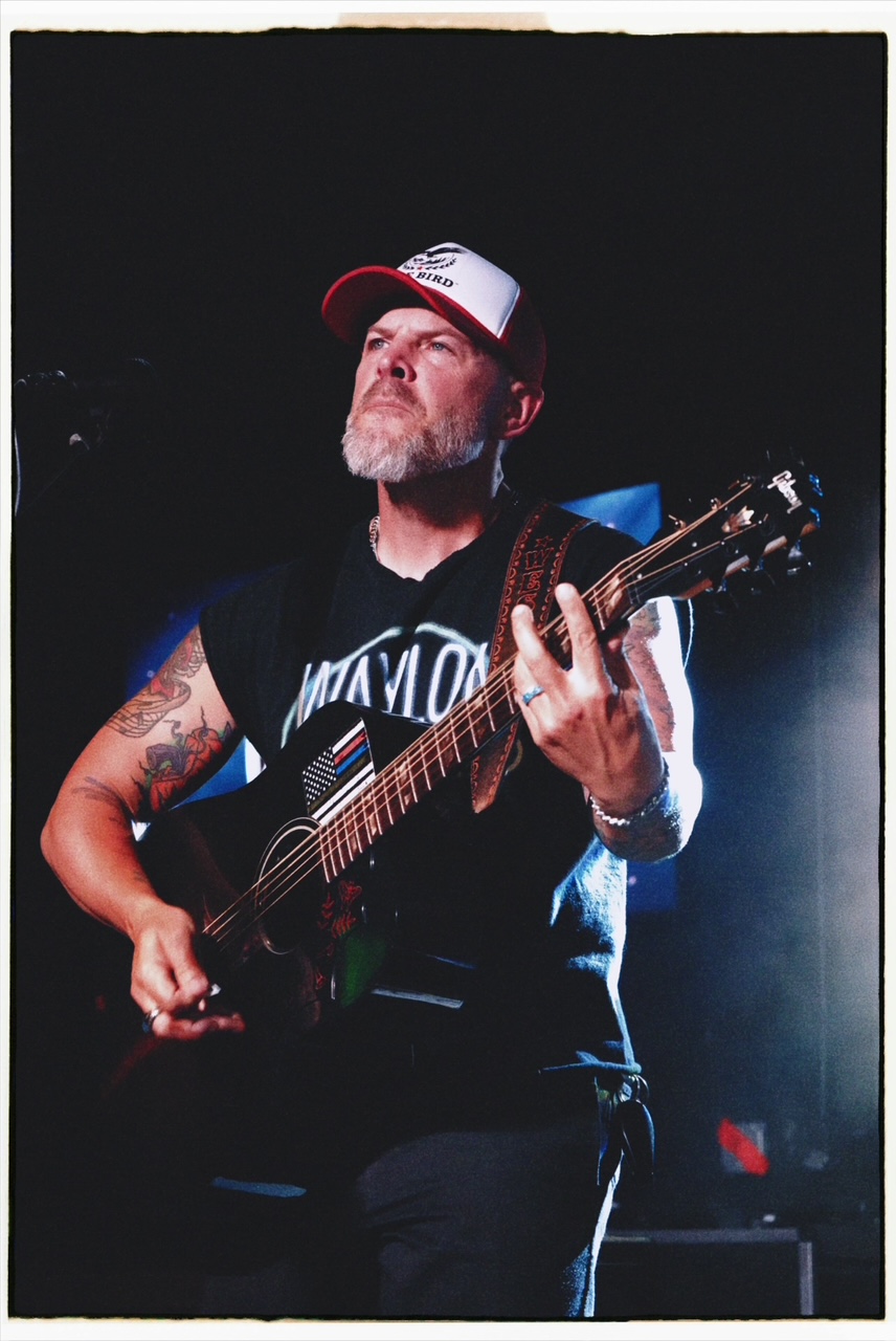 Man with a beard and tattoos playing an acoustic guitar, wearing a cap and sleeveless shirt, on stage with dark background.