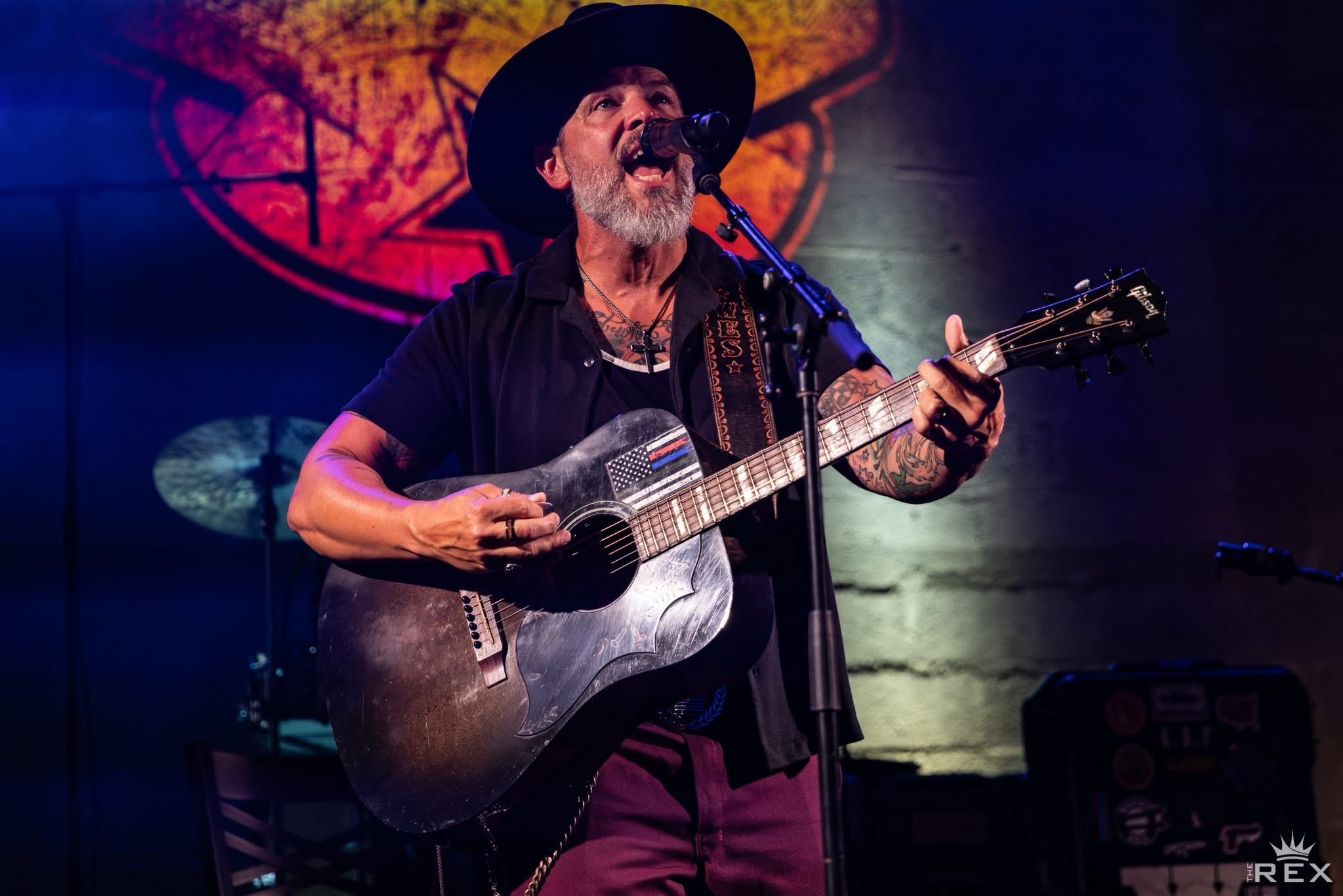 Man with a beard and hat playing an acoustic guitar on stage, singing into a microphone, with colorful background lighting.