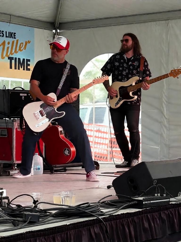 Two musicians playing electric guitars on stage inside a tent, with equipment and a sign in the background.
