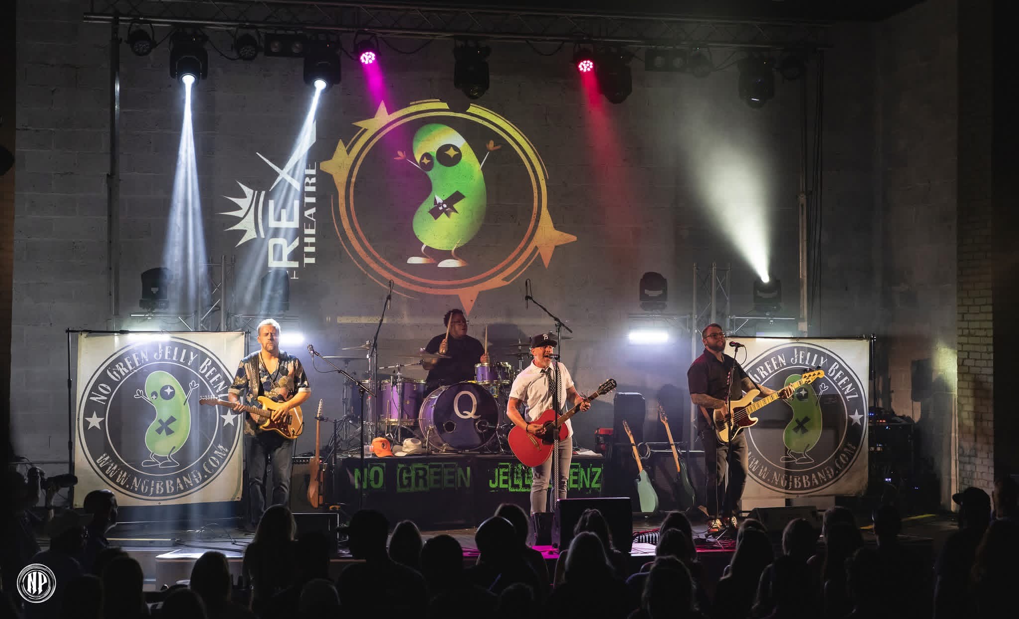 Band performing on stage with a large circular logo featuring a green character, stage lights, and audience silhouettes.