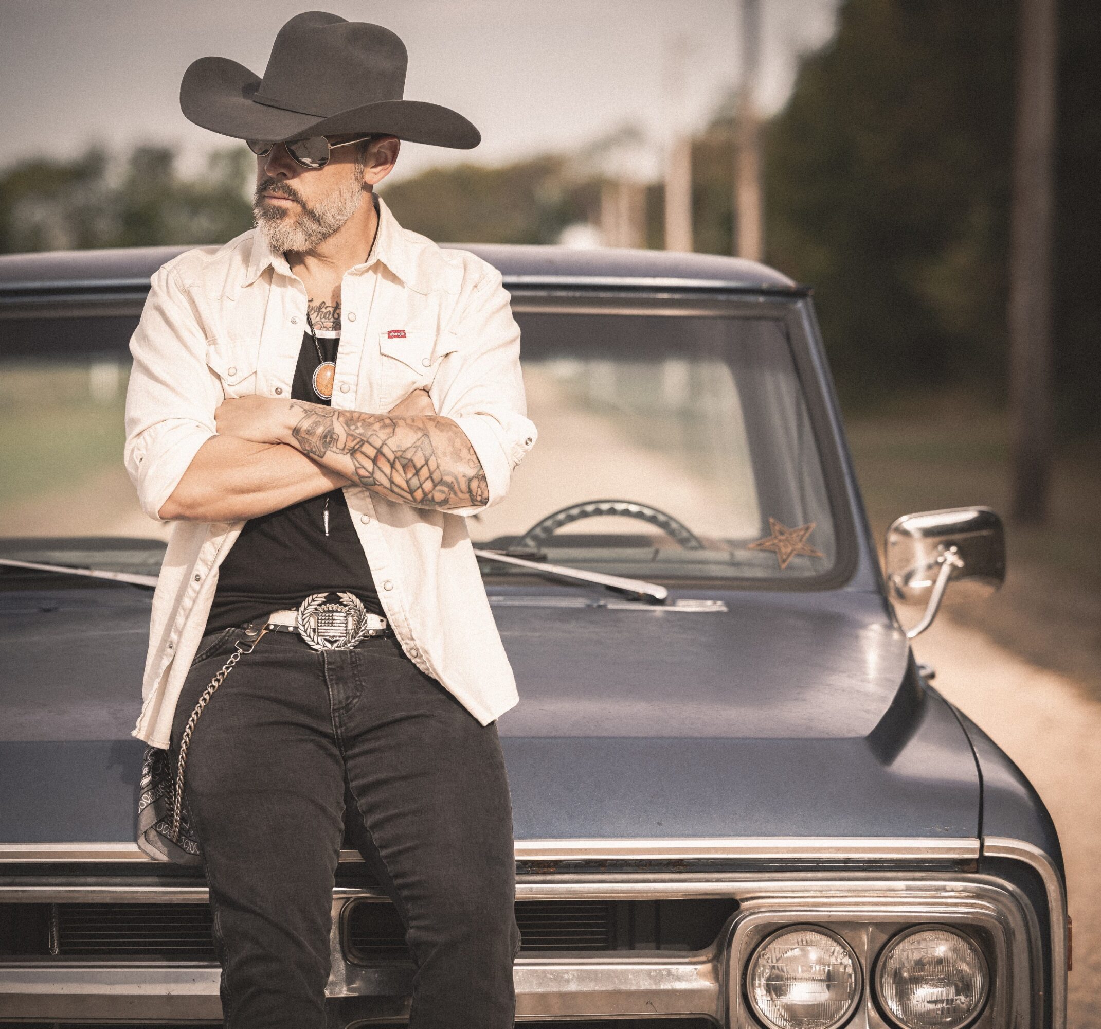 Man with a large hat and sunglasses leaning against a black vehicle, arms crossed, outdoors during daytime.