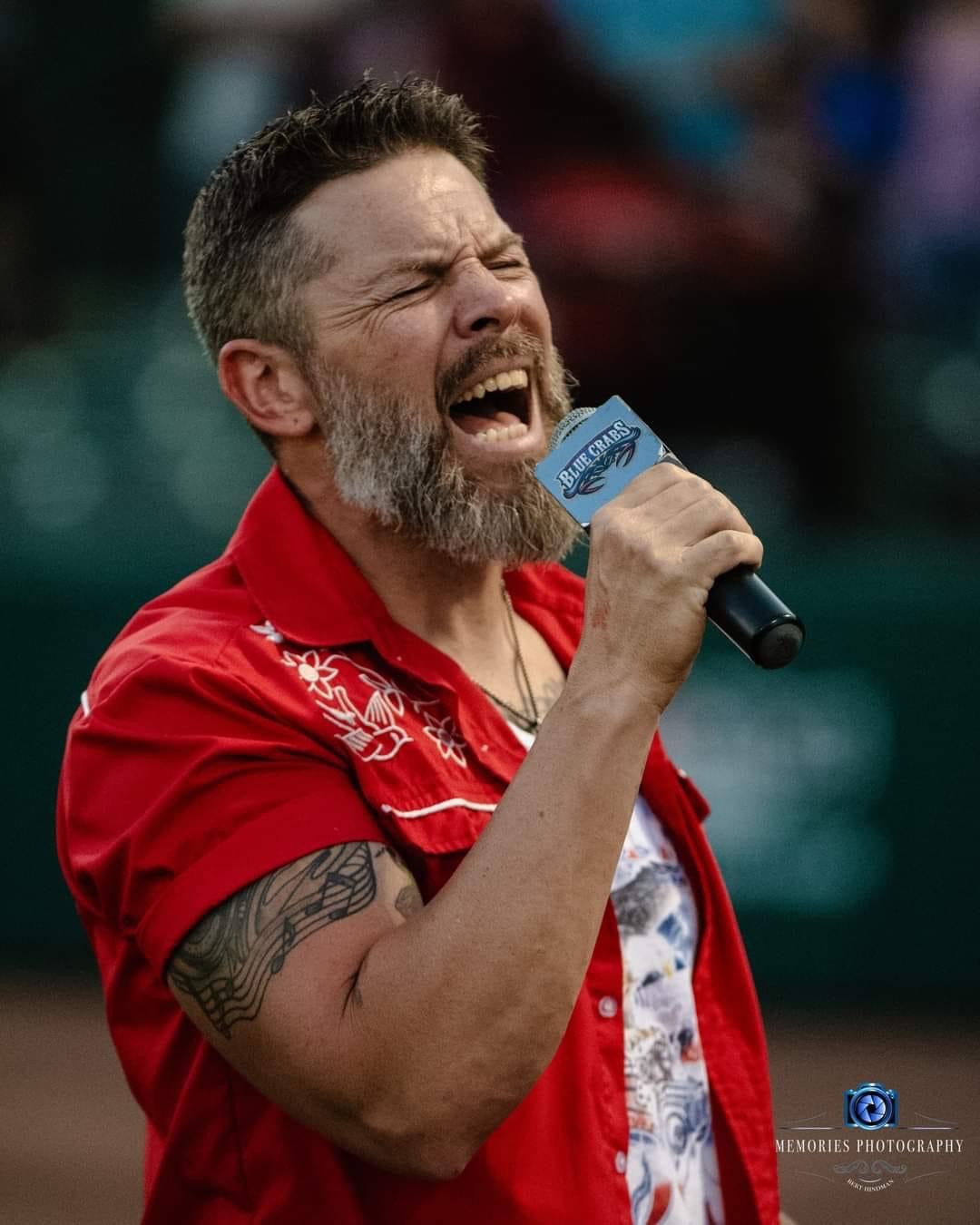Man with a beard and tattoos singing into a microphone outdoors, wearing a red shirt with white embroidery.