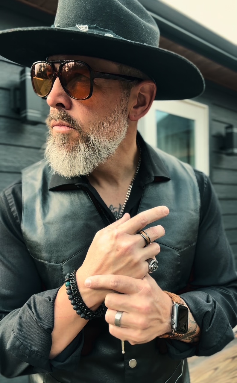Man with a beard wearing sunglasses, a wide-brimmed hat, and a leather vest, standing outdoors with a building in the background.