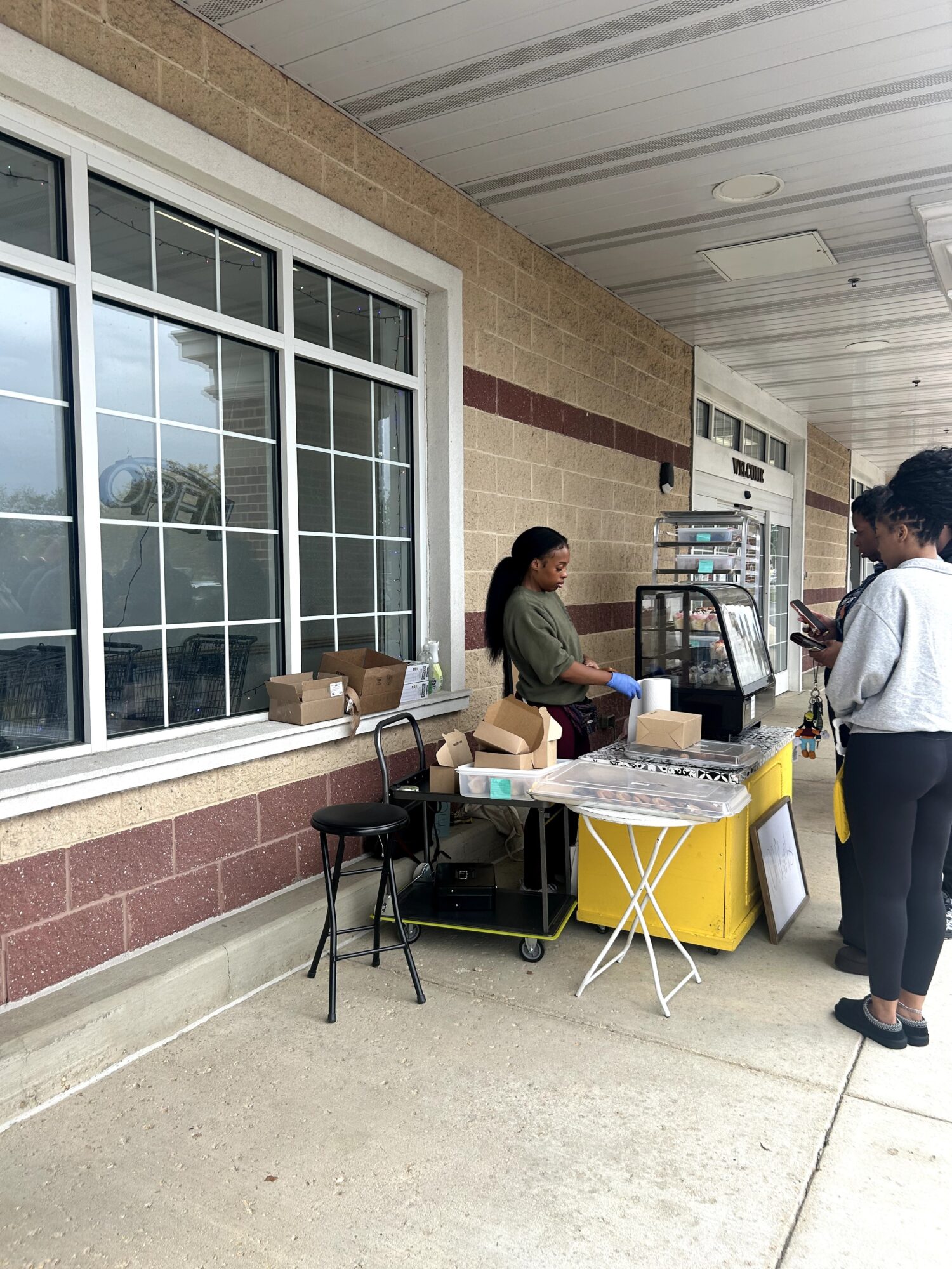 Two women at a small outdoor stand with a table, boxes, and a yellow cooler, near a large window and vending machines.