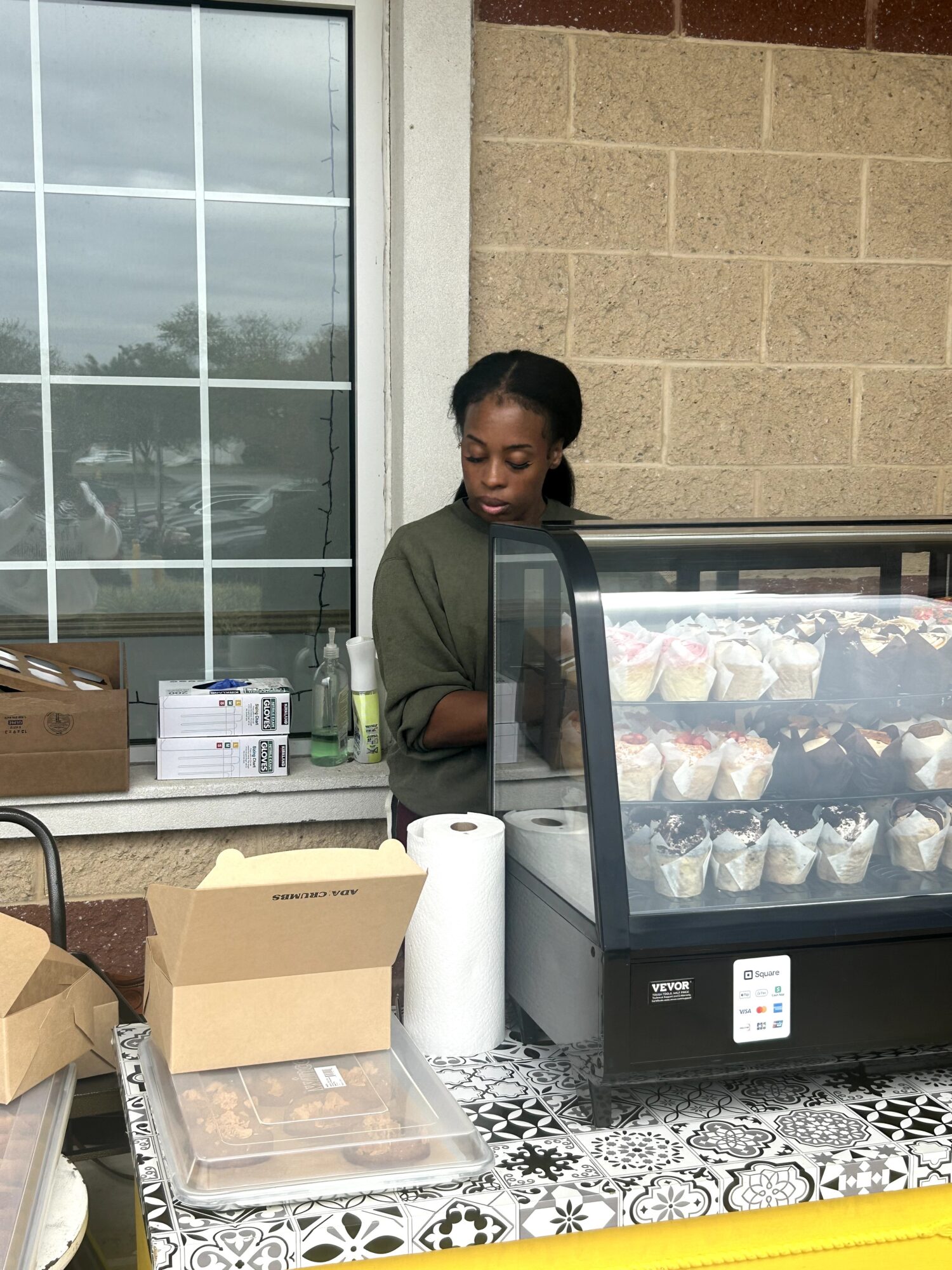 Young woman standing behind a display case with desserts, on a patterned table, outside a building with a window.