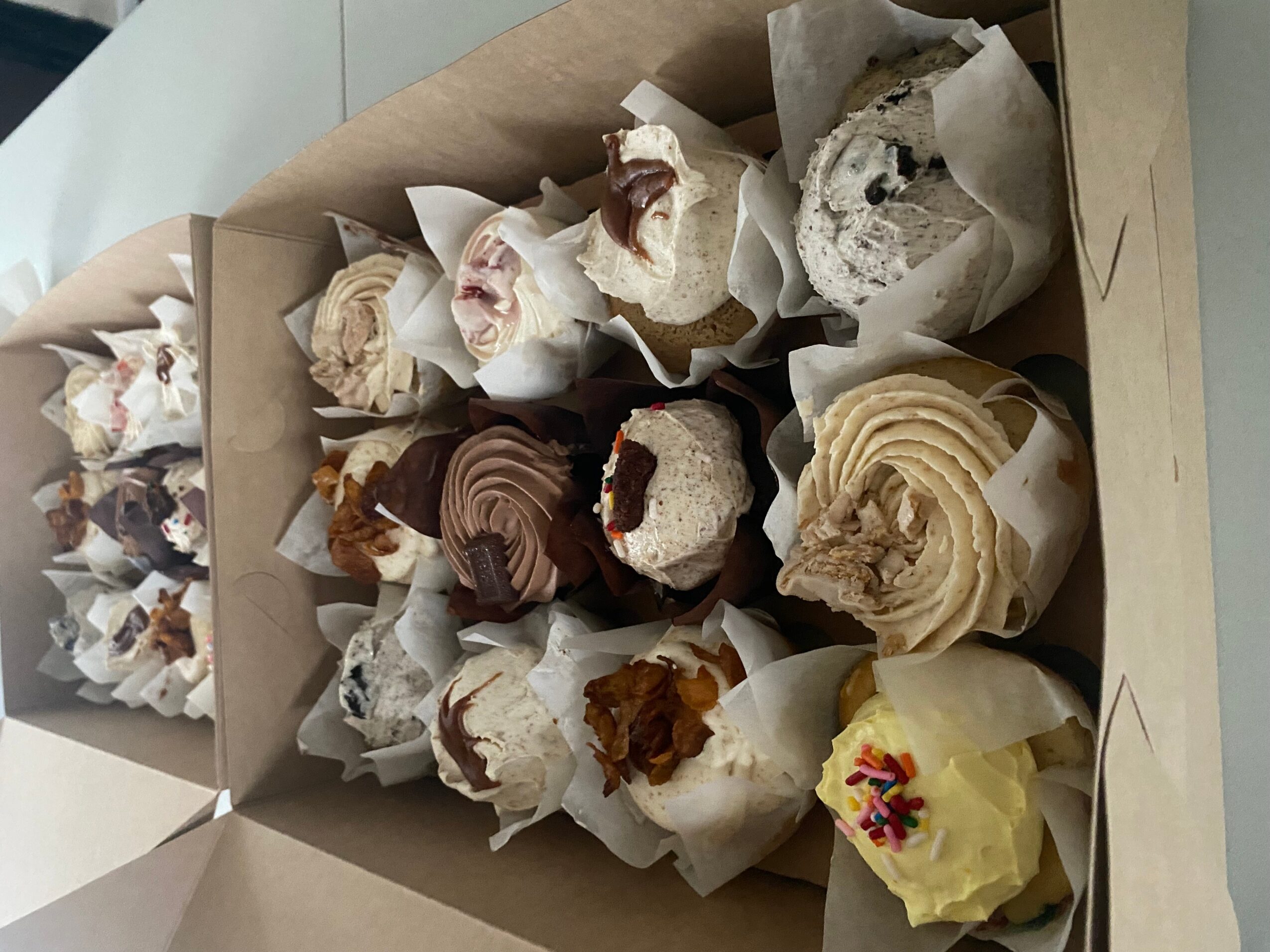 Box of assorted cupcakes with various frosting and toppings, some with sprinkles and others with chocolate or fruit decorations.