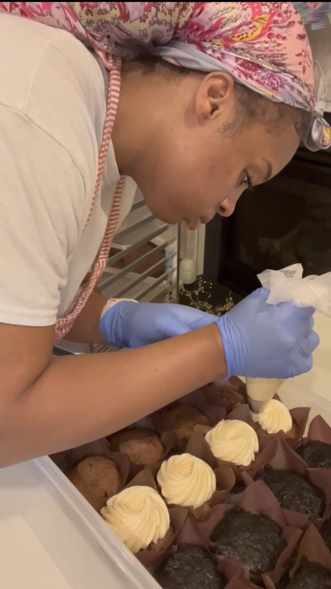 Person wearing a headscarf and gloves decorating baked goods with frosting, focusing on the task.