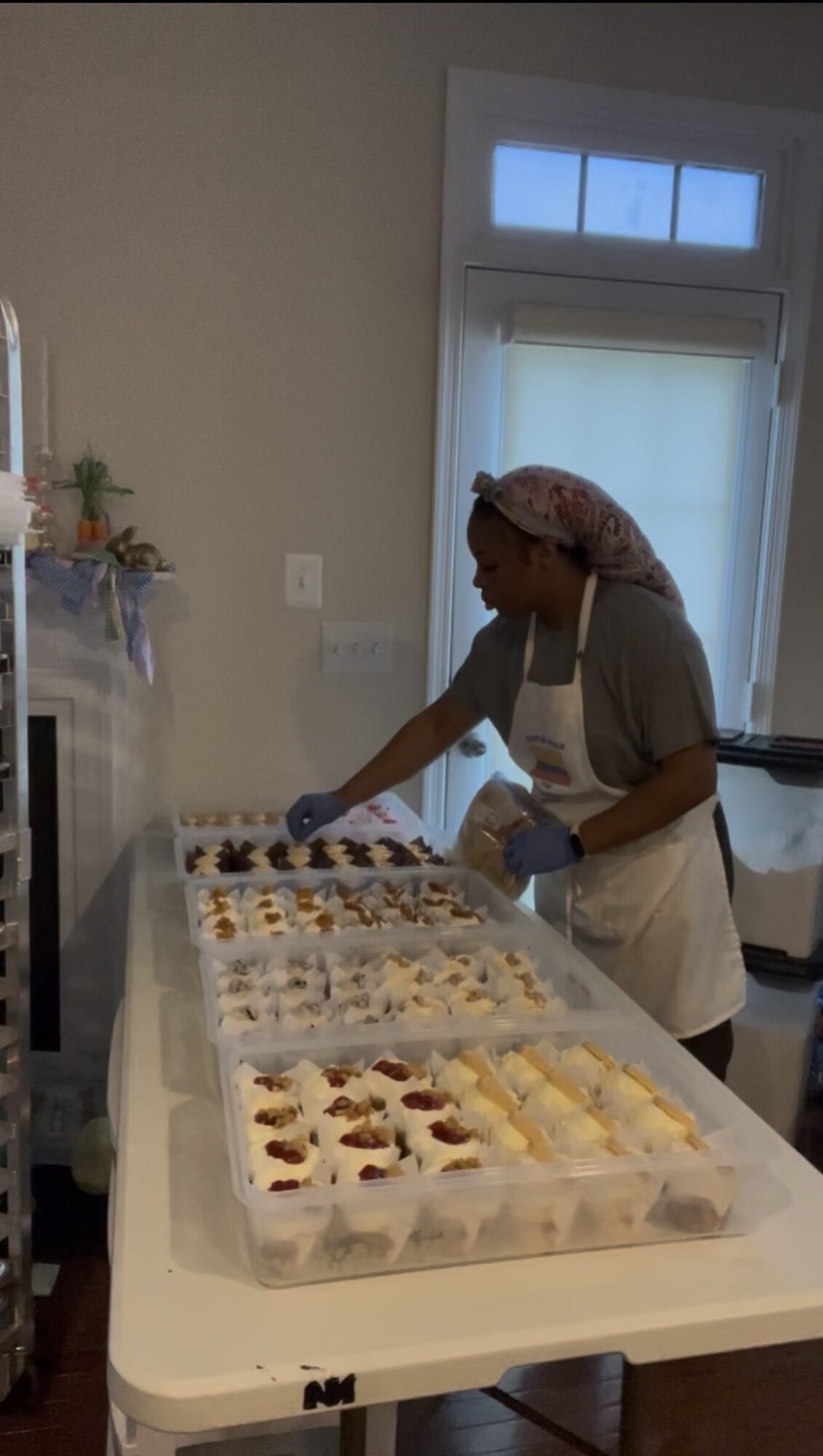 Person wearing a headscarf and apron arranging desserts on a table in a bright room.