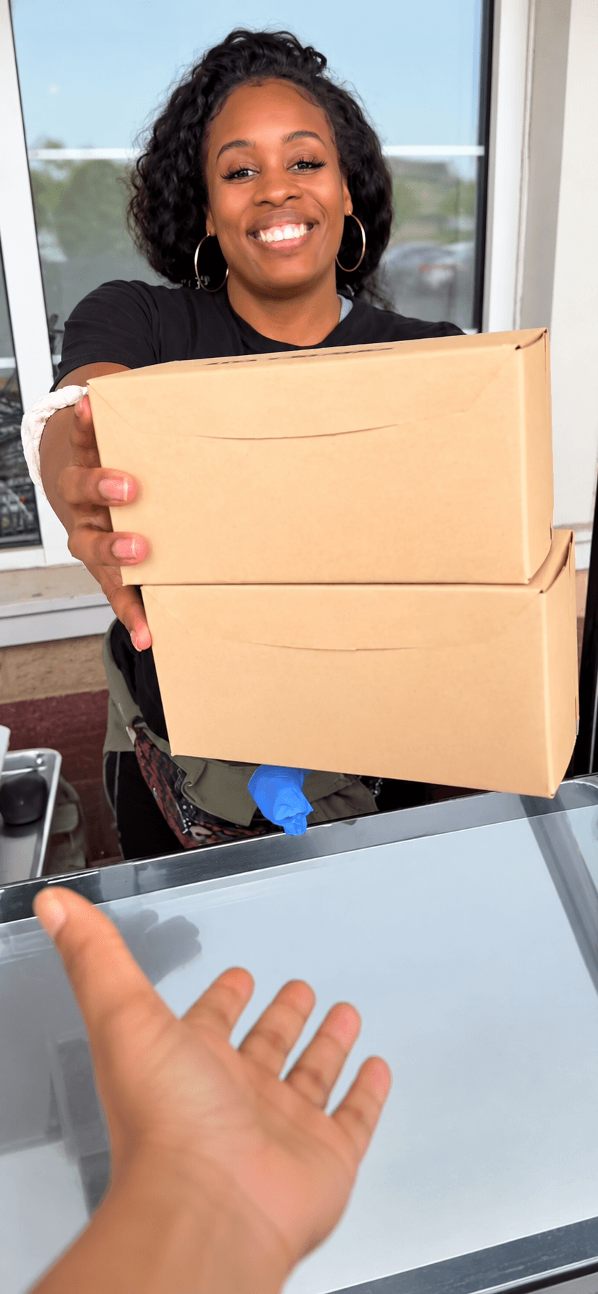 Person smiling and holding two cardboard boxes, standing indoors near a window.