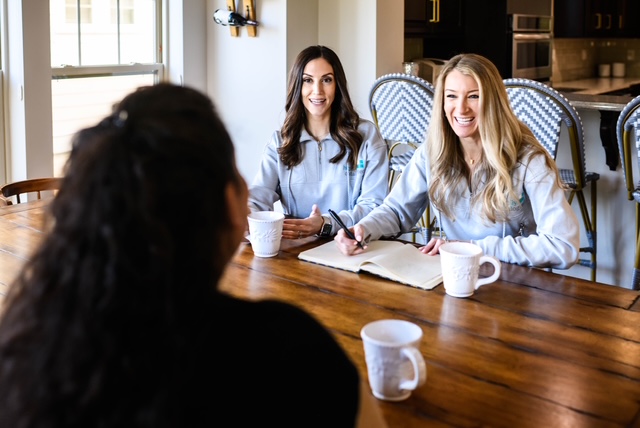 Three women sit at a wooden table in a bright room, engaging in conversation with notebooks and mugs in front of them.