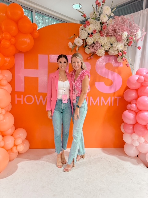 Two women stand in front of a pink and orange backdrop with balloons and flowers, smiling at the camera.
