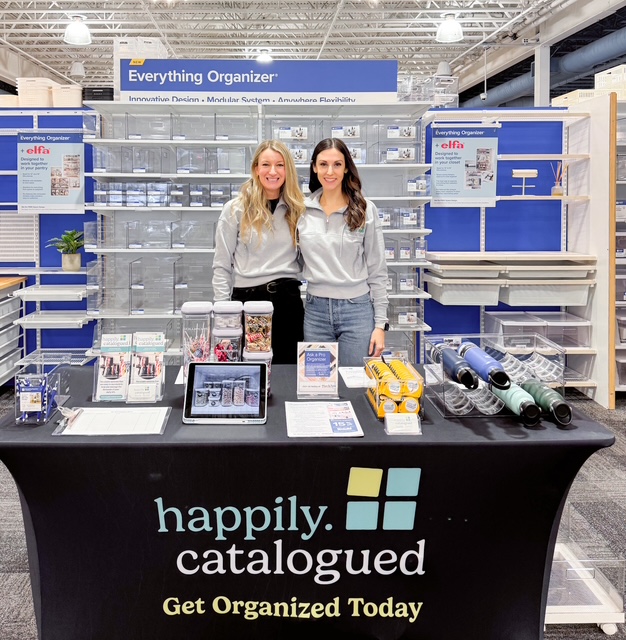 Two women stand behind a table with organized products in a store aisle, smiling at the camera.
