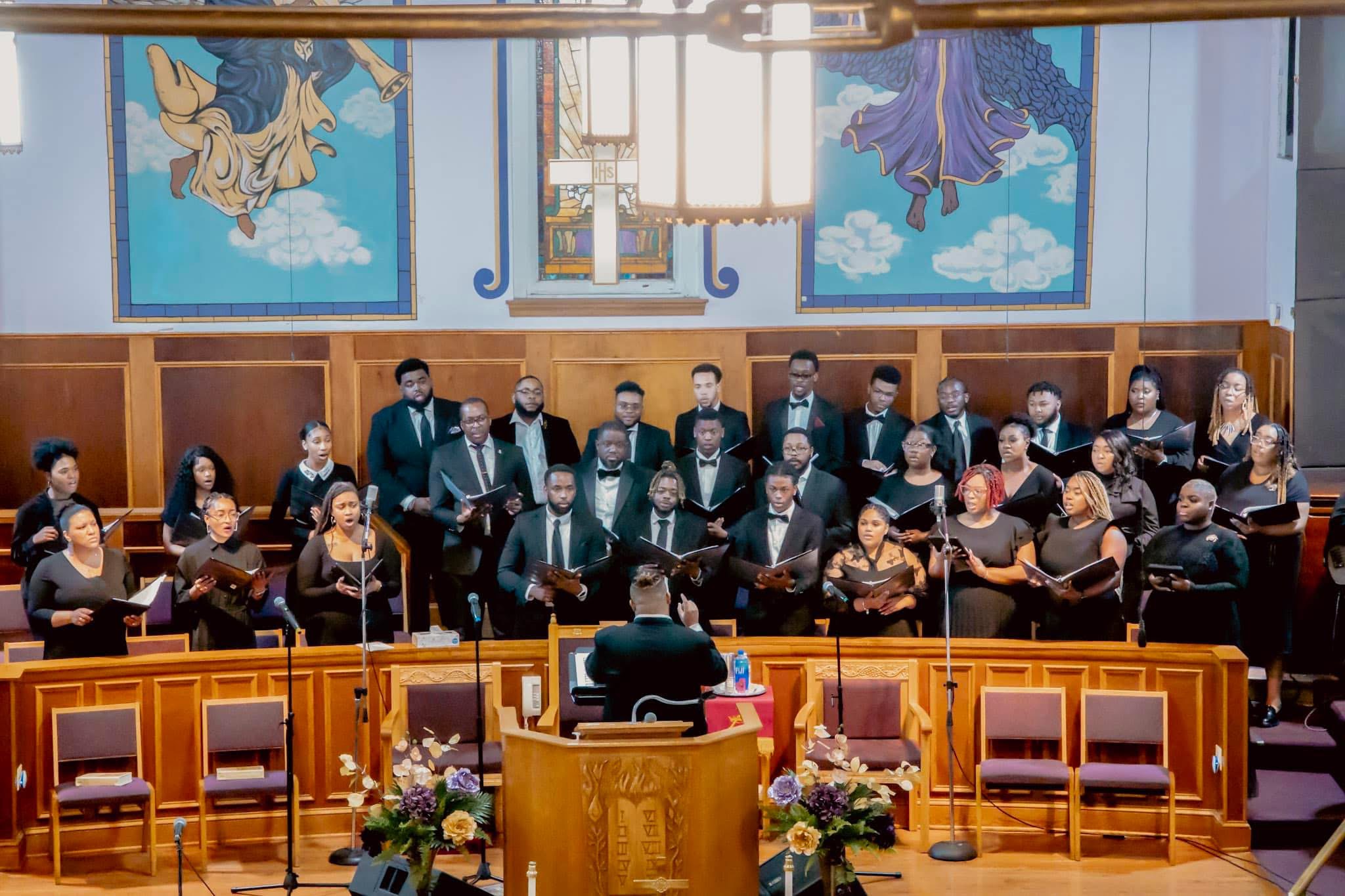Choir and musicians perform on stage in a church with stained glass windows, wooden paneling, and floral arrangements.