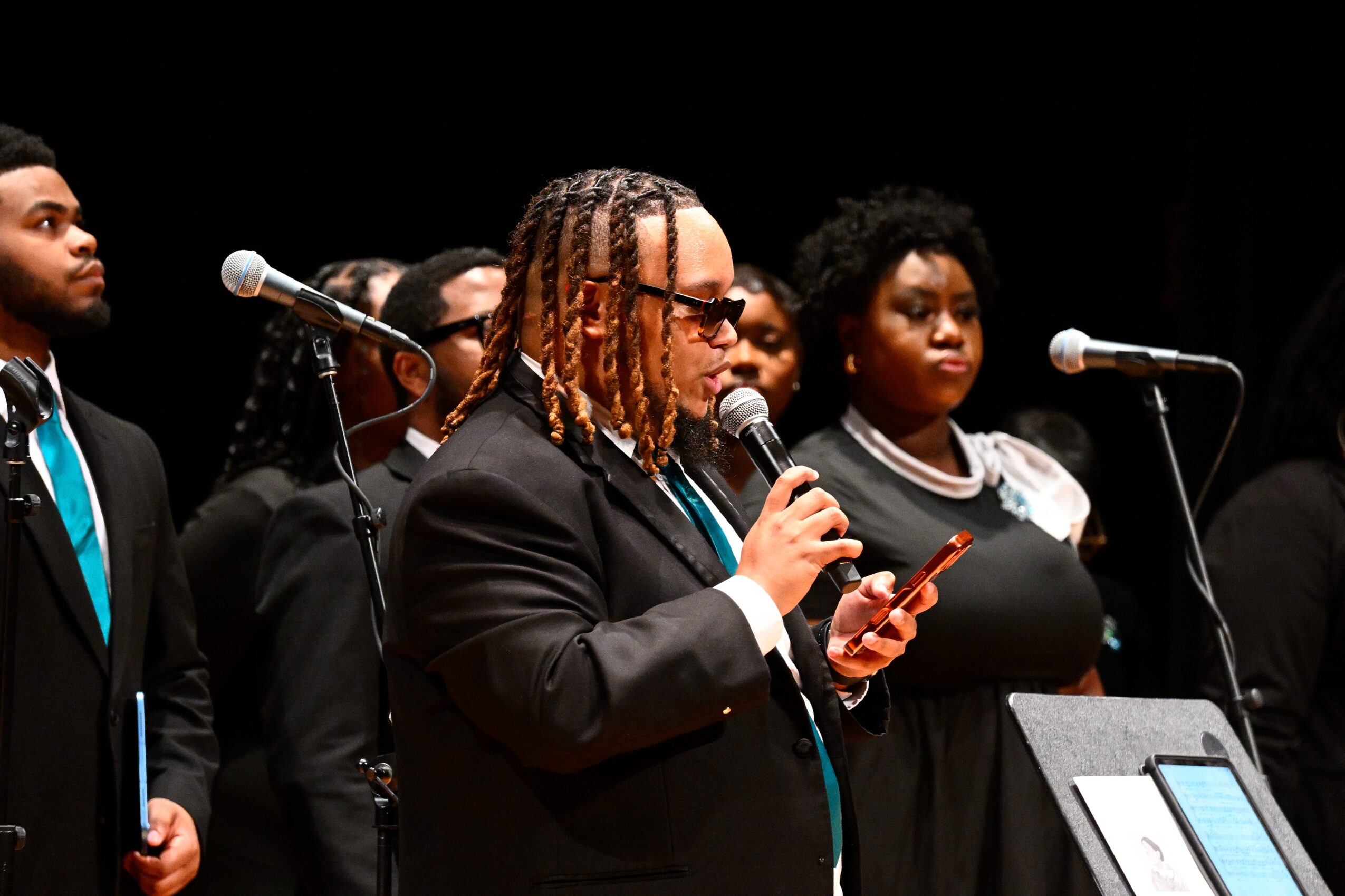 Group of people standing on stage, one person speaking into a microphone and reading from a phone.
