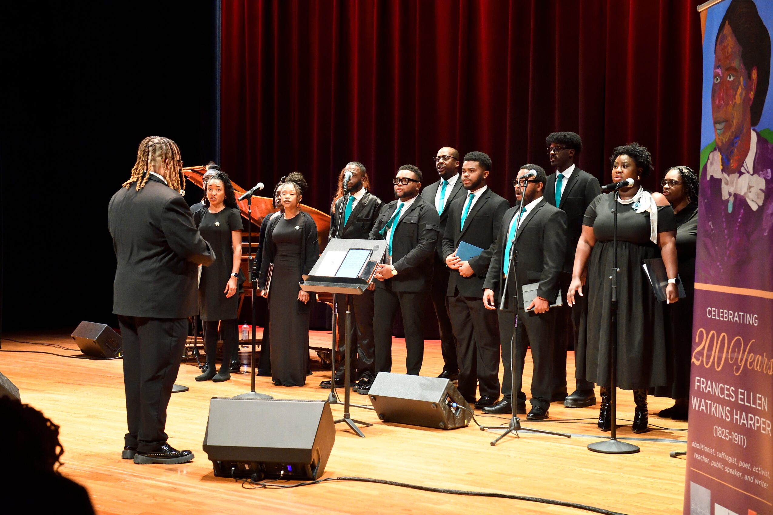 Choir of people singing on stage with a conductor, red curtain background, and a poster on the right side.