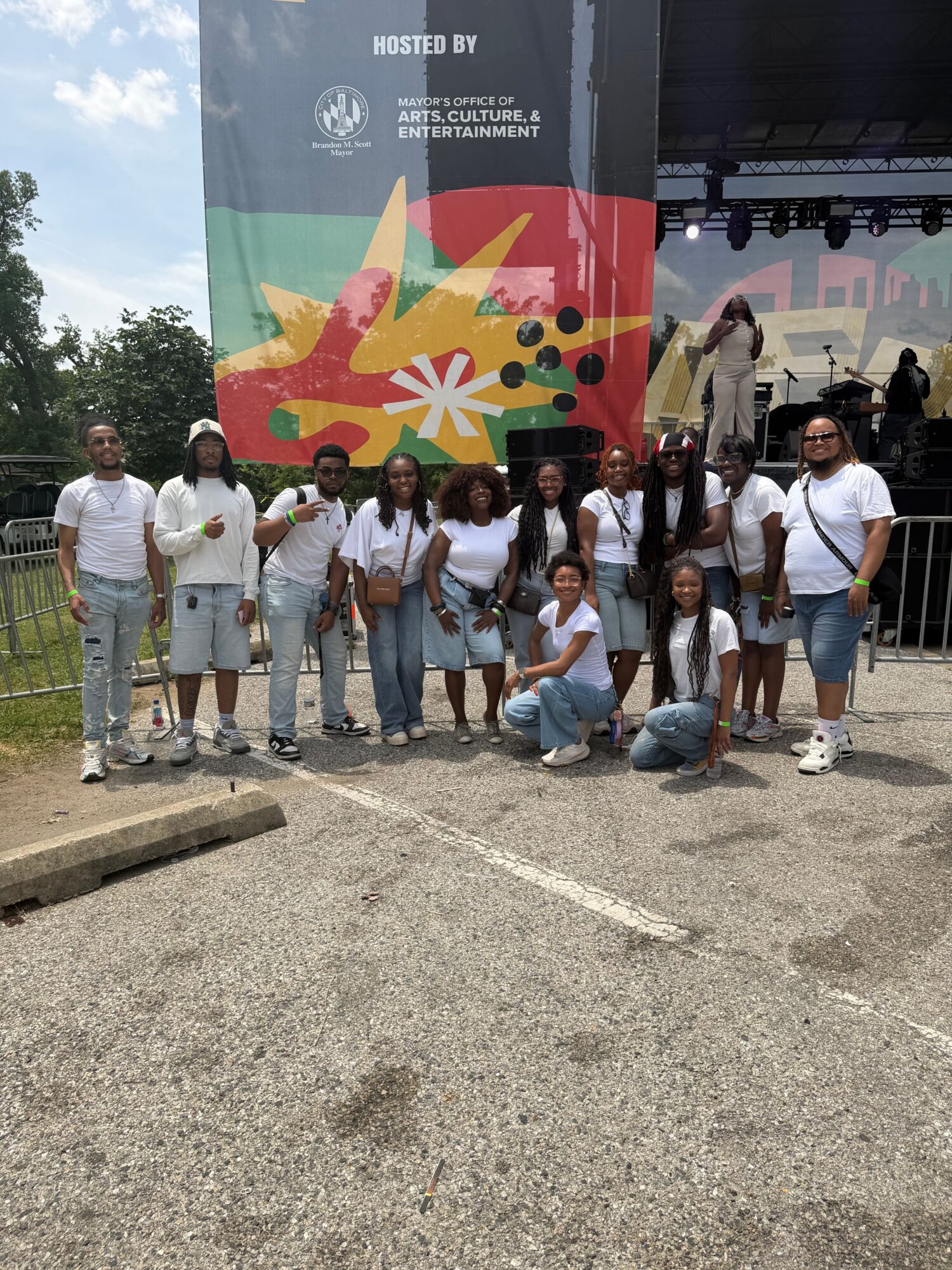 Group of children and adults standing outdoors in front of a colorful banner, some holding bags, on a sunny day.