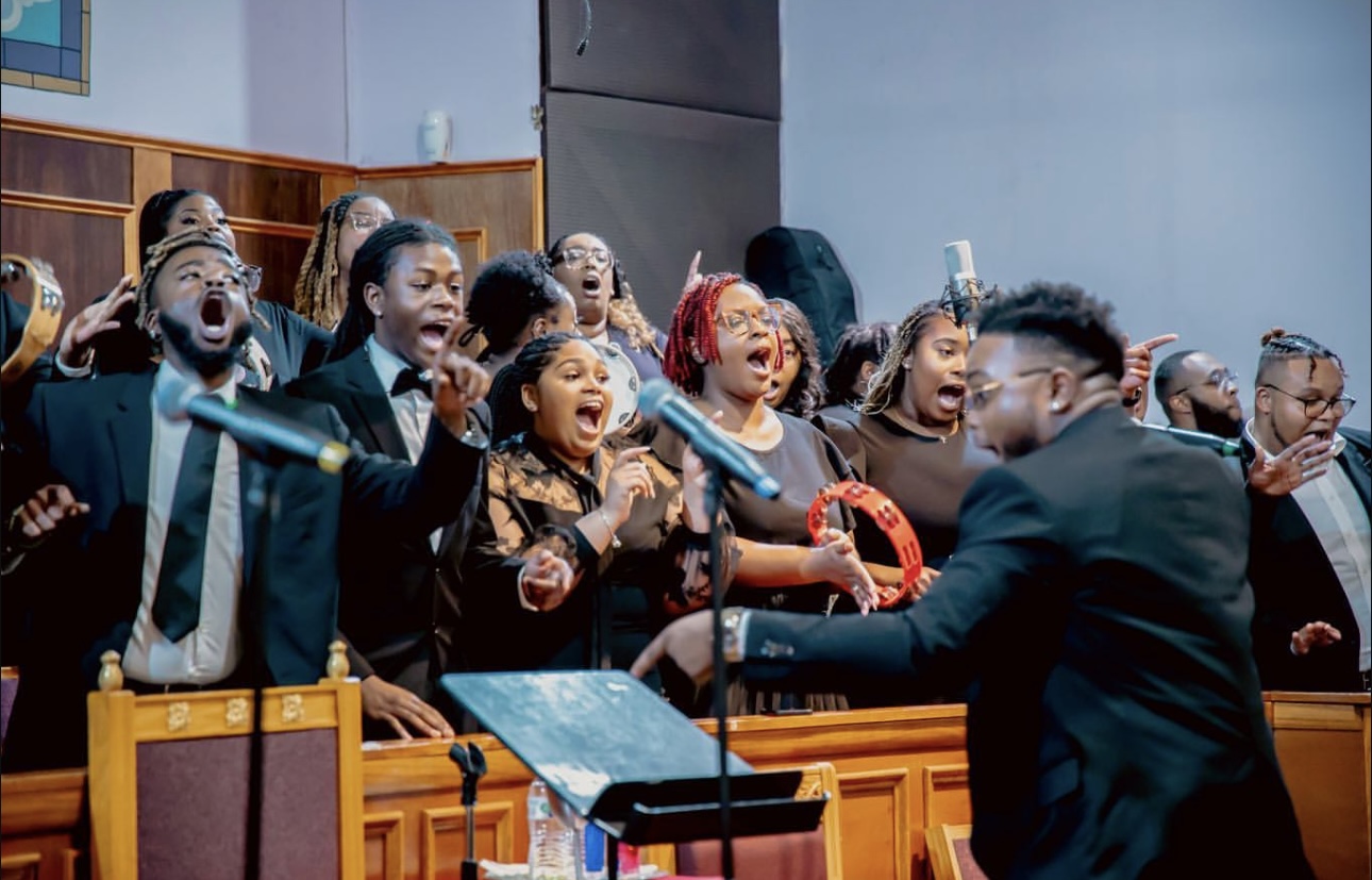 Children singing passionately in a choir, led by a conductor, in a church or auditorium setting.