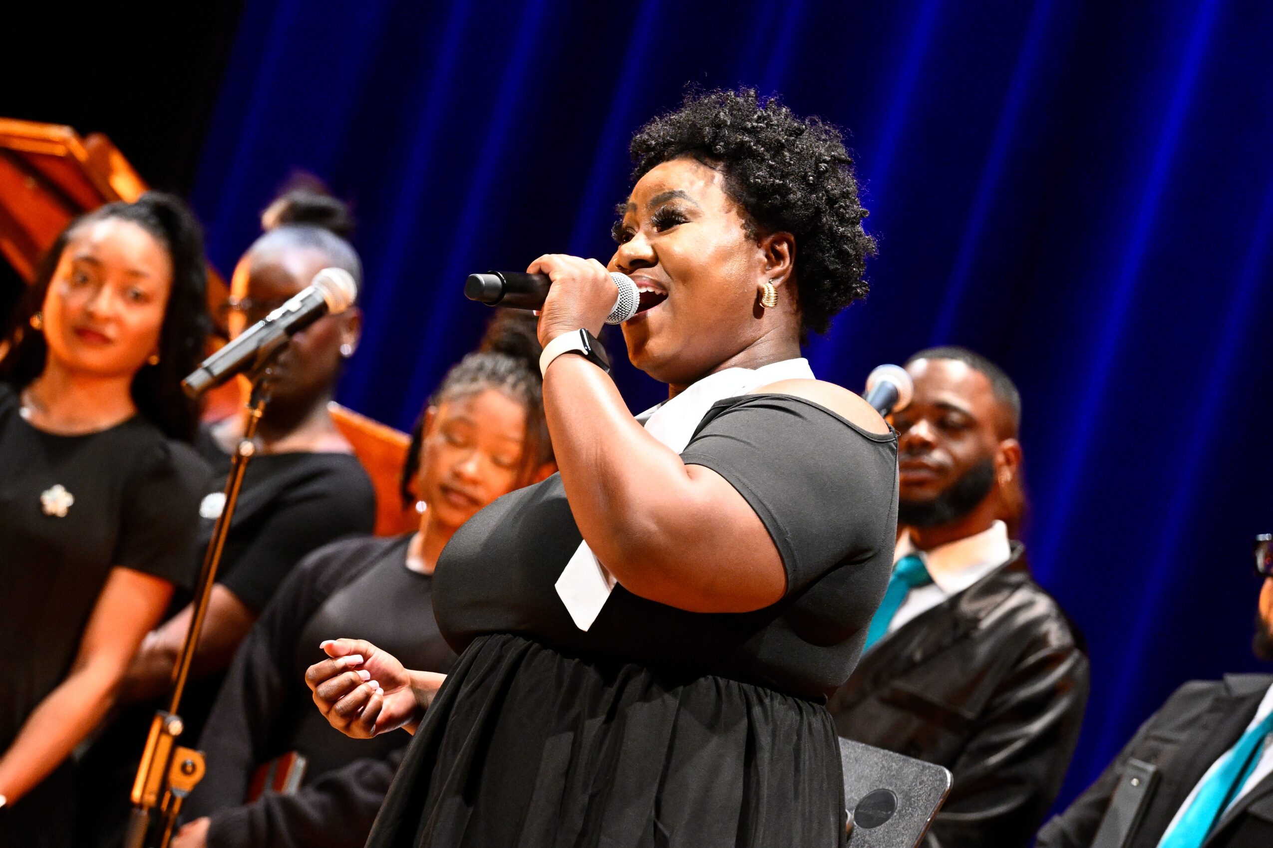 Group of people singing on stage, woman in front holding a microphone, blue curtain background.