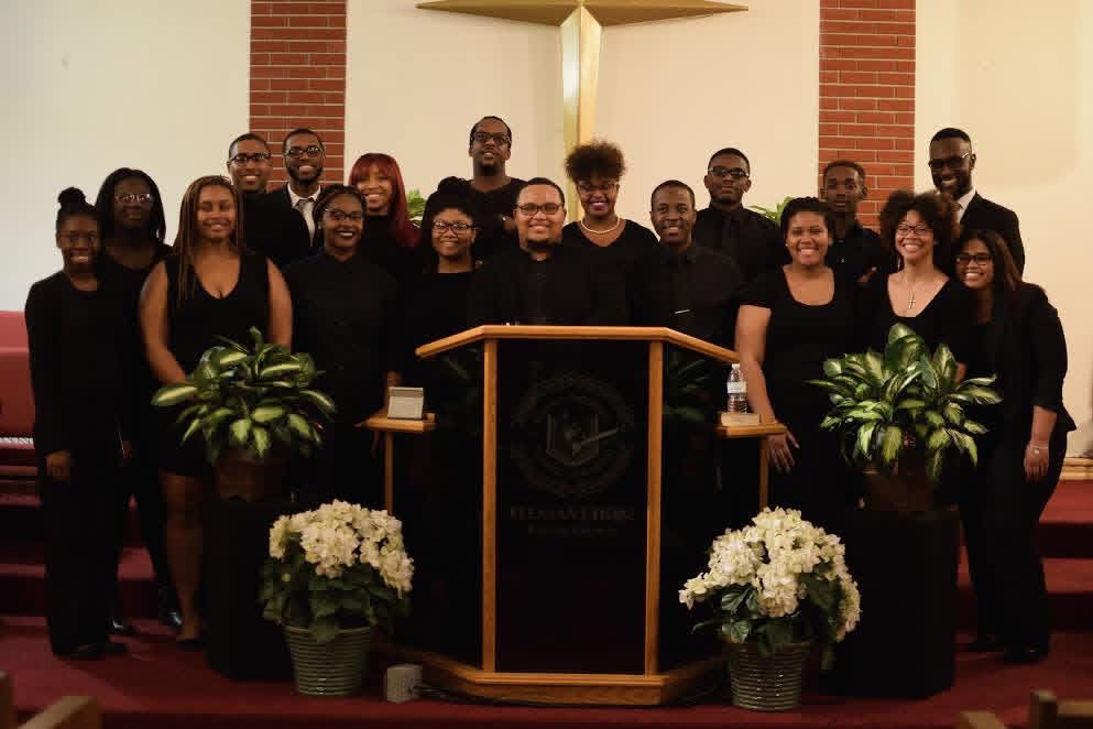 Group of people standing on stage behind a podium with flowers, in a church or auditorium setting.