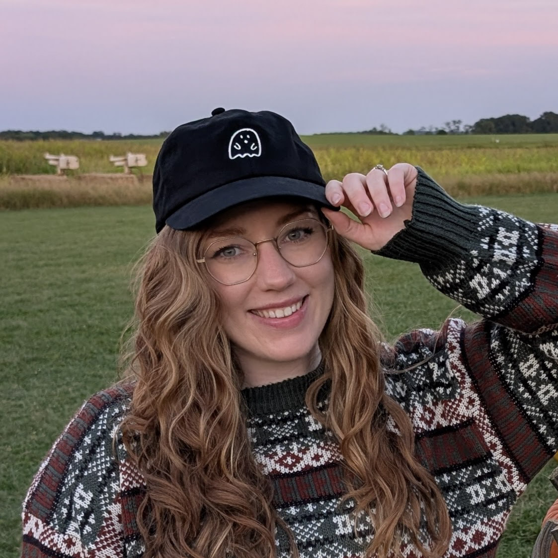 Woman with long wavy hair wearing glasses, a patterned sweater, and a black cap outdoors, smiling at the camera.