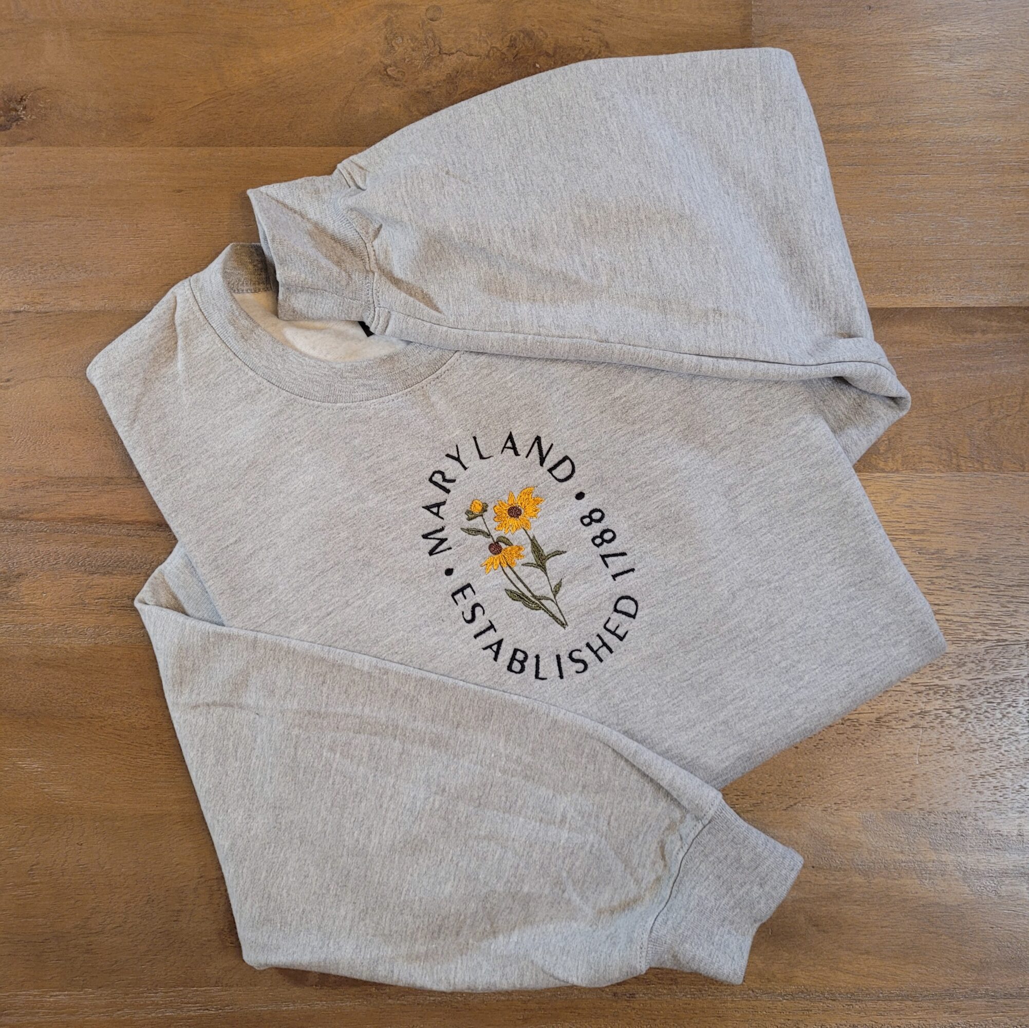 Gray sweatshirt with a sunflower graphic and text, laid on a wooden surface.