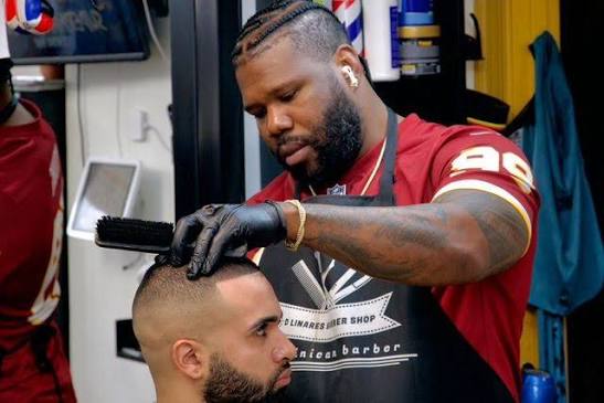 Barber trims a man's beard with a brush in a barbershop, both focused on grooming.