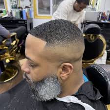 Man with a fresh fade haircut and gray beard sitting in a barbershop, with barber working in background.