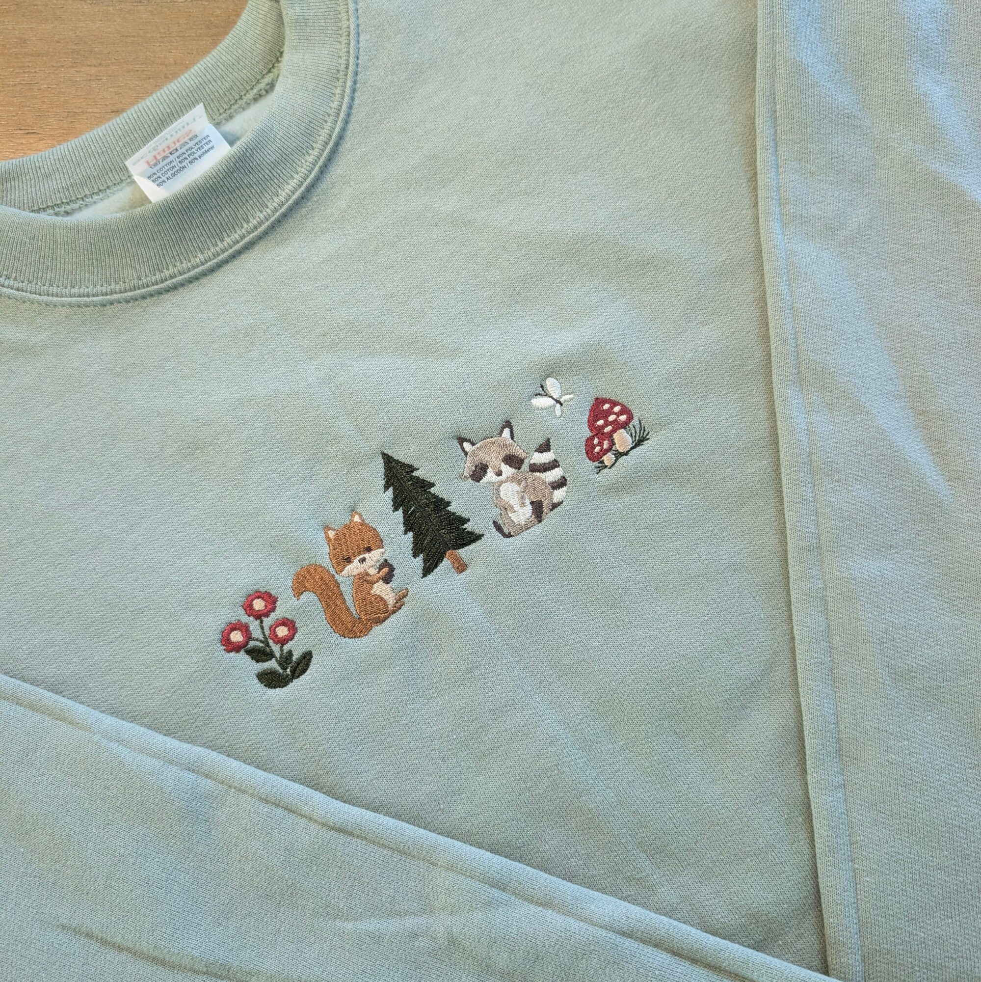 Light green shirt with embroidered fox, tree, raccoon, mushroom, and flowers design on chest area.